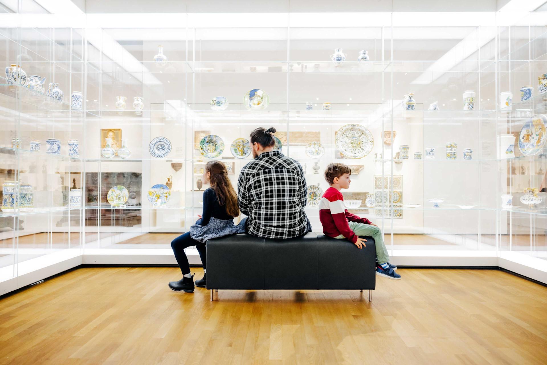 Un père et ses deux enfants sont assis sur un tabouret dans une salle d’œuvres d’art.