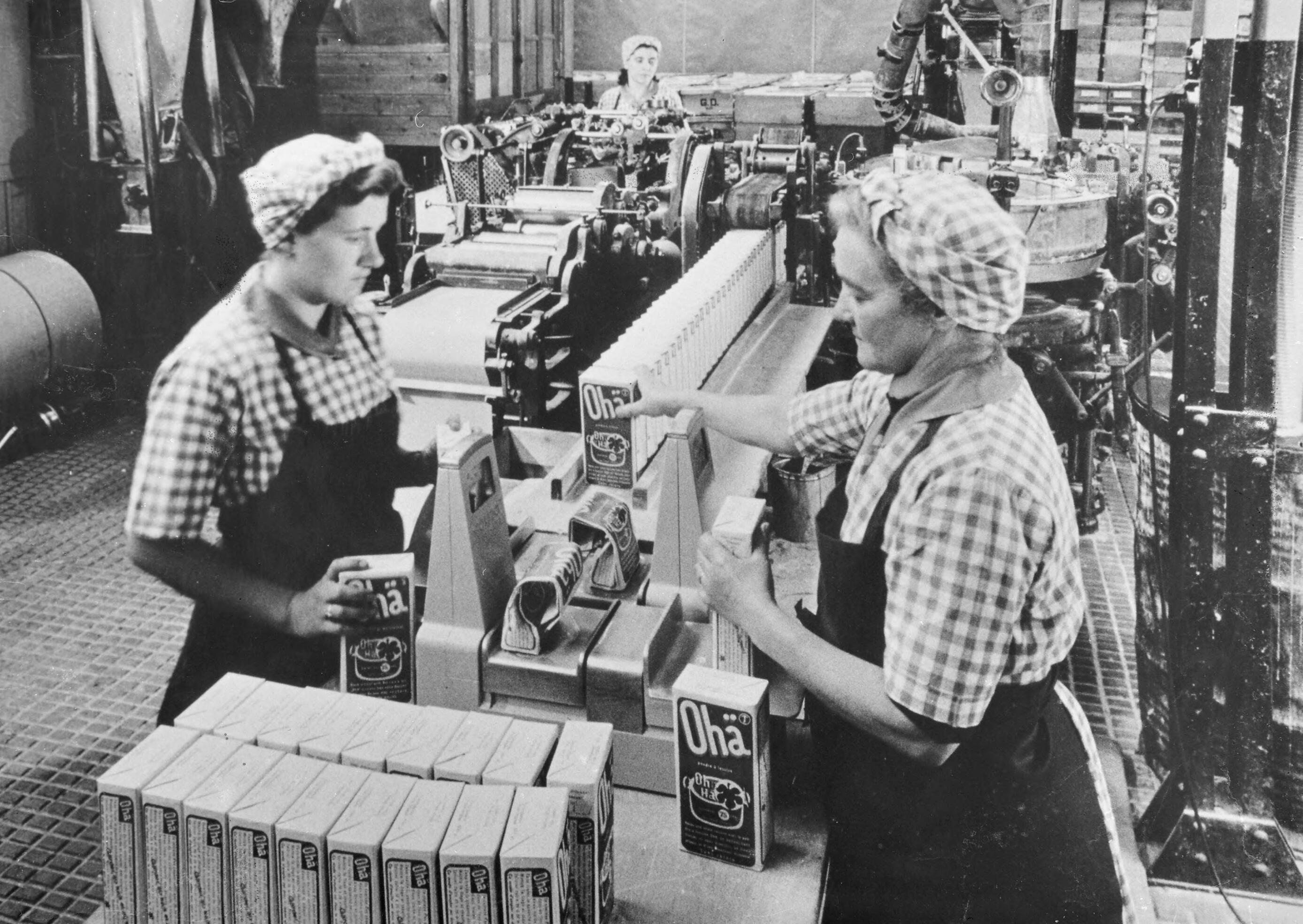 A black-and-white photograph of women packing Ohä on a conveyor belt.