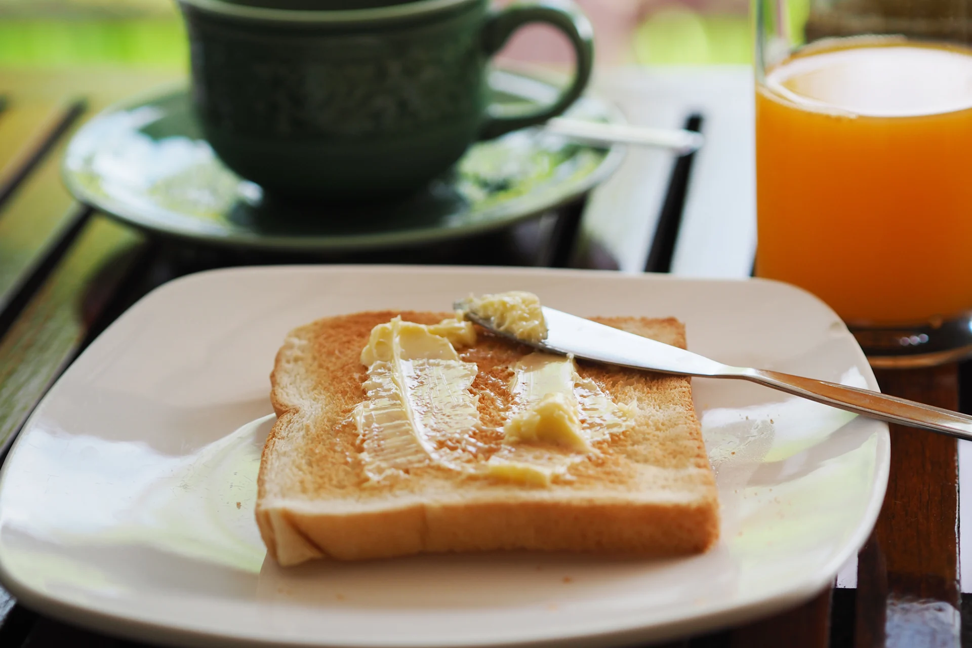 A buttered piece of toast on a plate. Behind it there is some orange juice and a cup containing a hot drink.