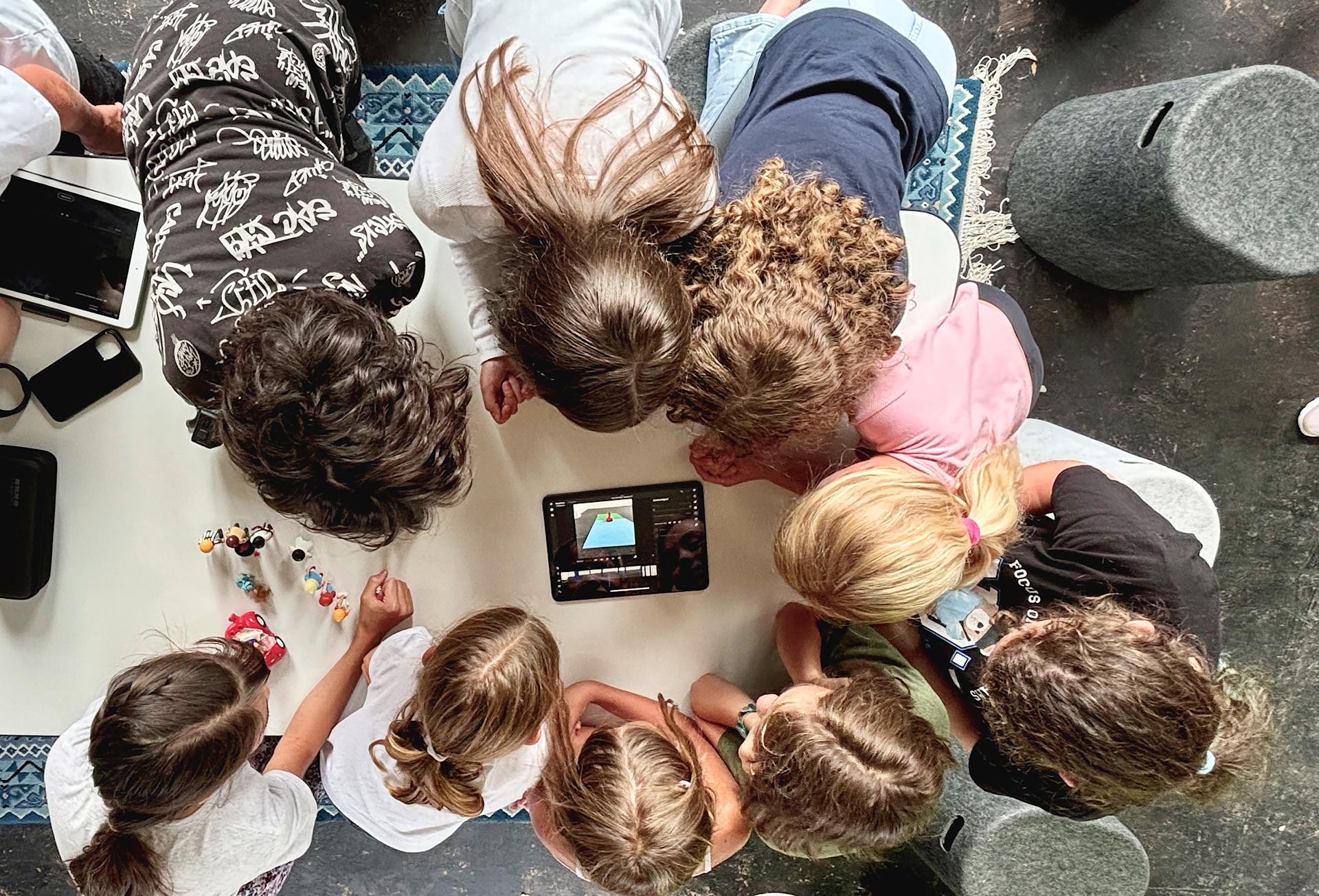 Plusieurs enfants sont assis autour d’une table, tête contre tête, les yeux rivés sur une tablette posée au milieu.