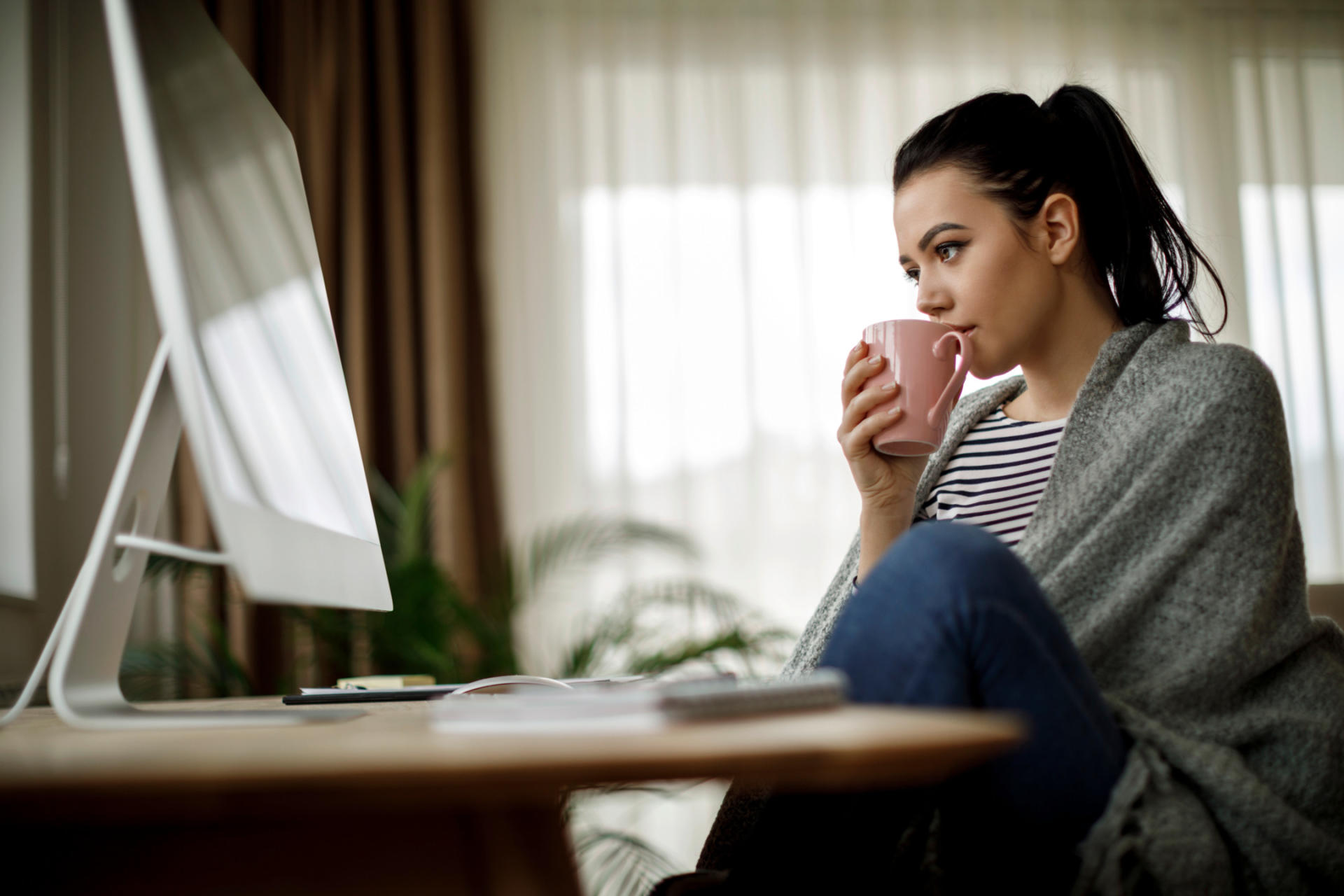 A woman sits in front of the computer with a cup of tea, wrapped in a blanket
