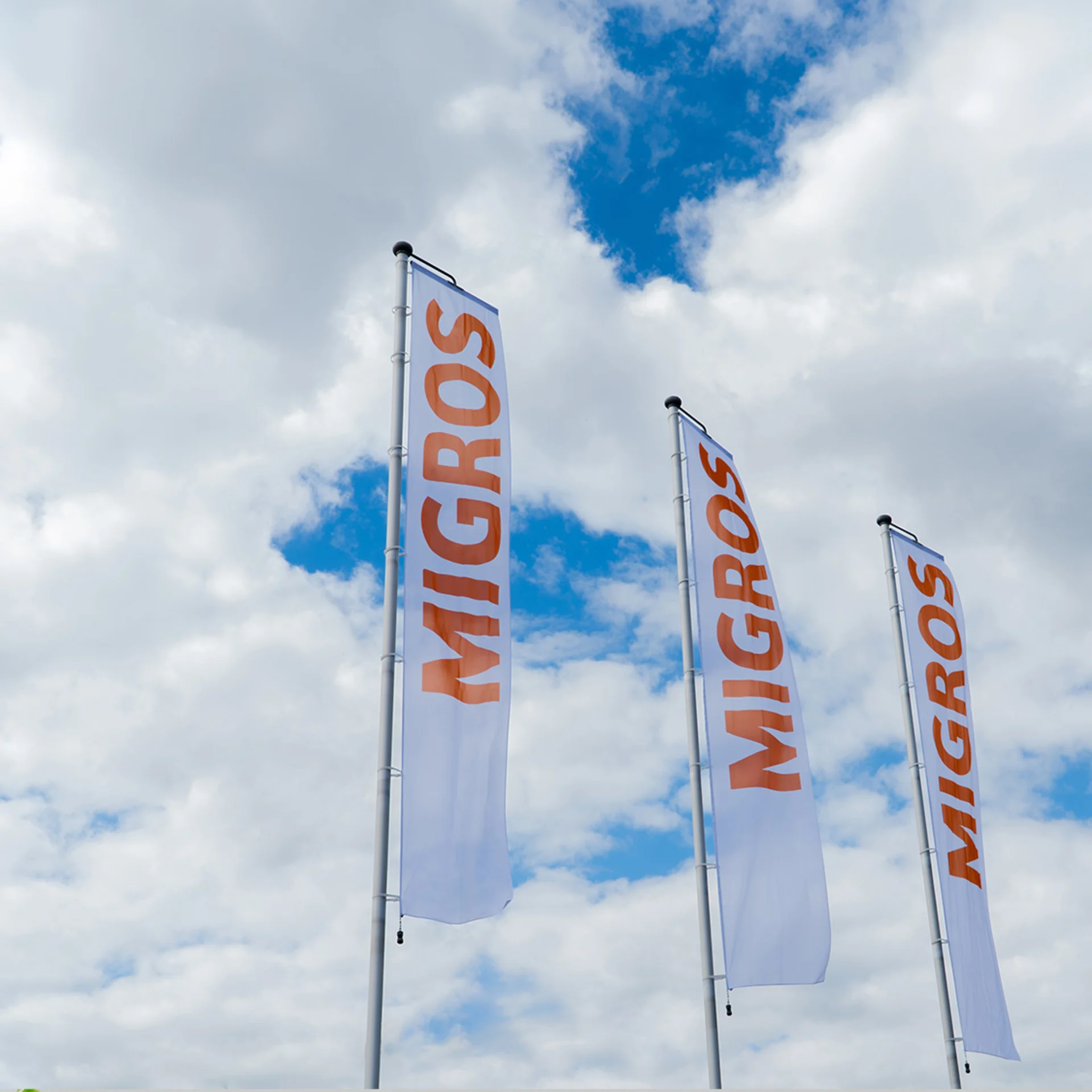3 oriflammes Migros sur fond de ciel nuageux