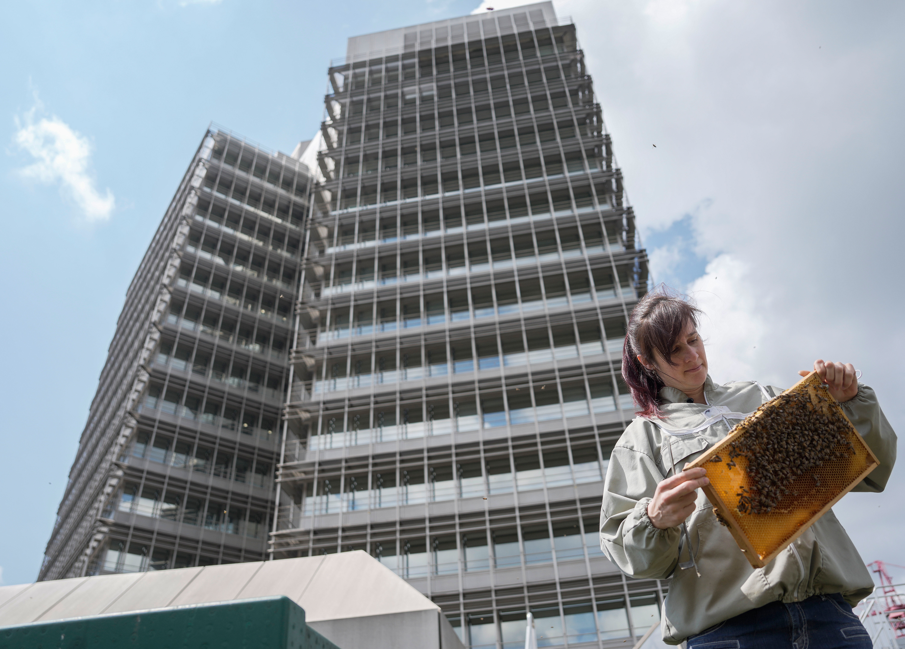 Mirjam Lendenmann ist auf dem Migros-Dach und betrachtet eine Bienenwabe. Im Hintergrund sieht man das Doppelhochhaus des Migros-Genossenschafts-Bundes.