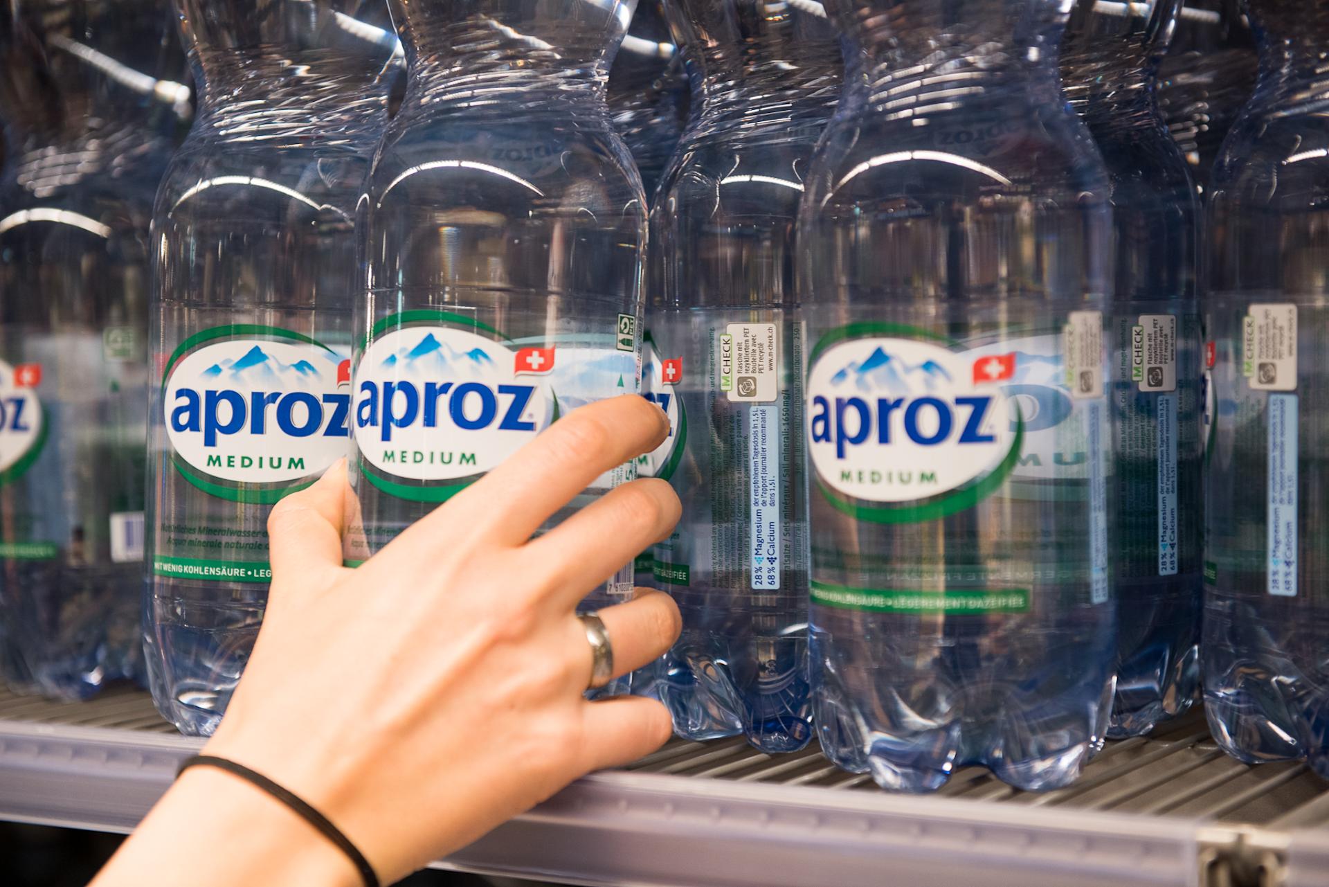 Une main saisissant une bouteille Aproz dans un rayon Migros.