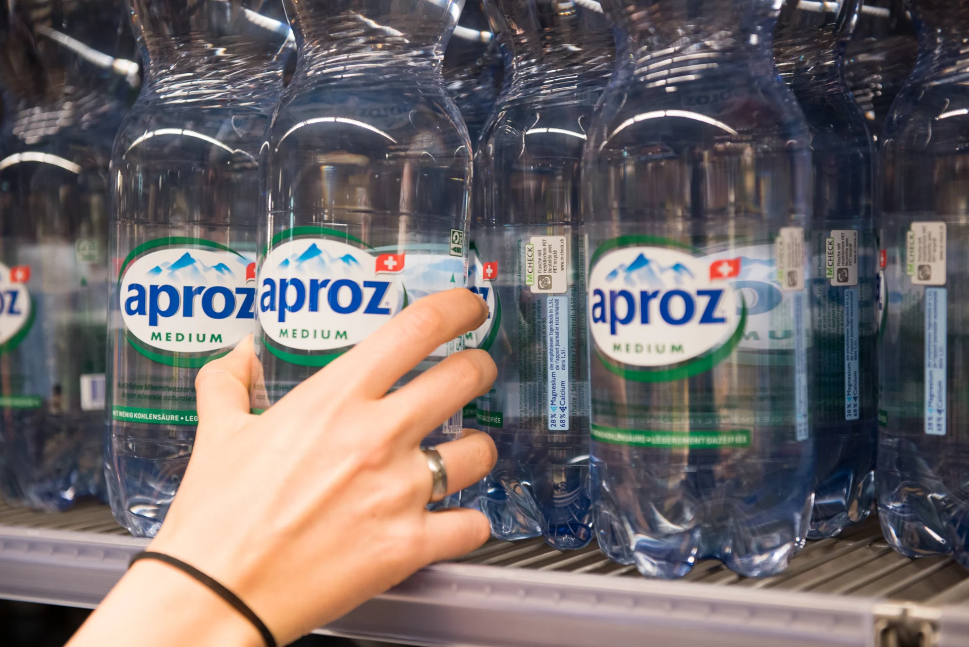 A hand reaches for an Aproz bottle on a shelf in Migros.