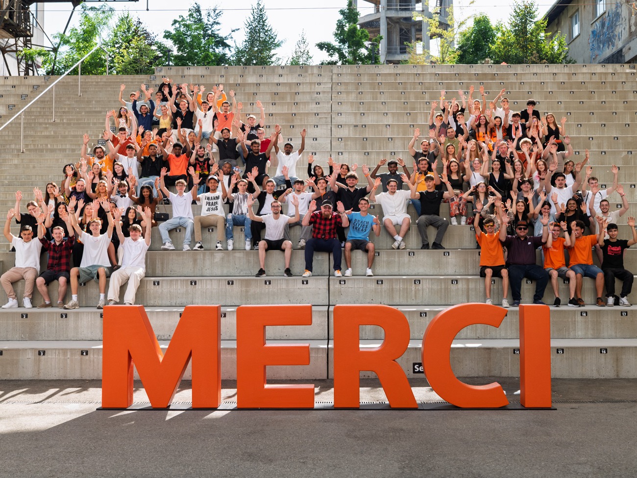 Grosse Gruppe junger Menschen sitzt und steht auf Steinstufen einer Aussenarena, alle mit erhobenen Armen, vor grossen MERCI-Buchstaben. 
