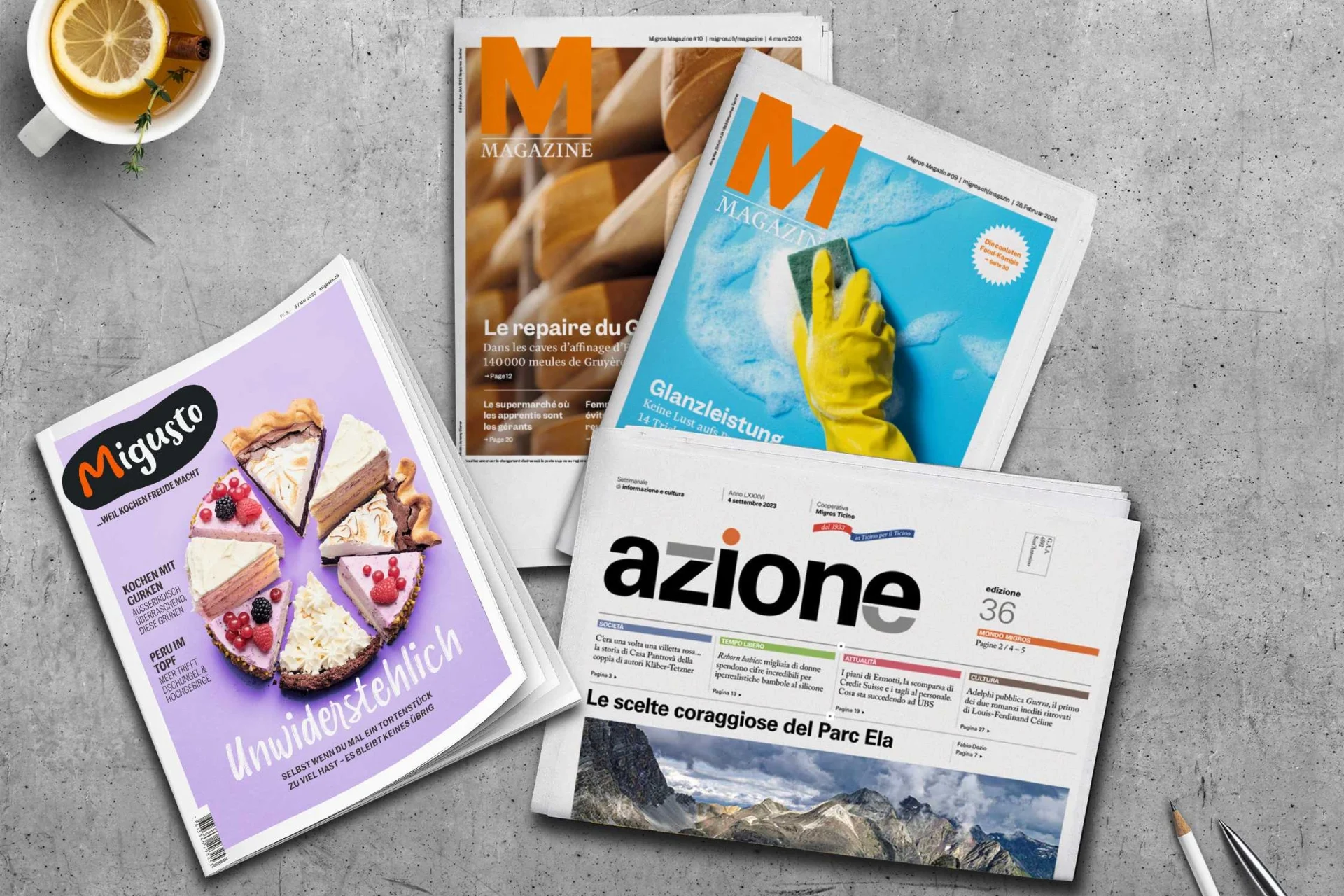 Ausgaben vom Migros Magazin, dem Migusto Kochmagazin und Azione liegen auf einem grauen Tisch.