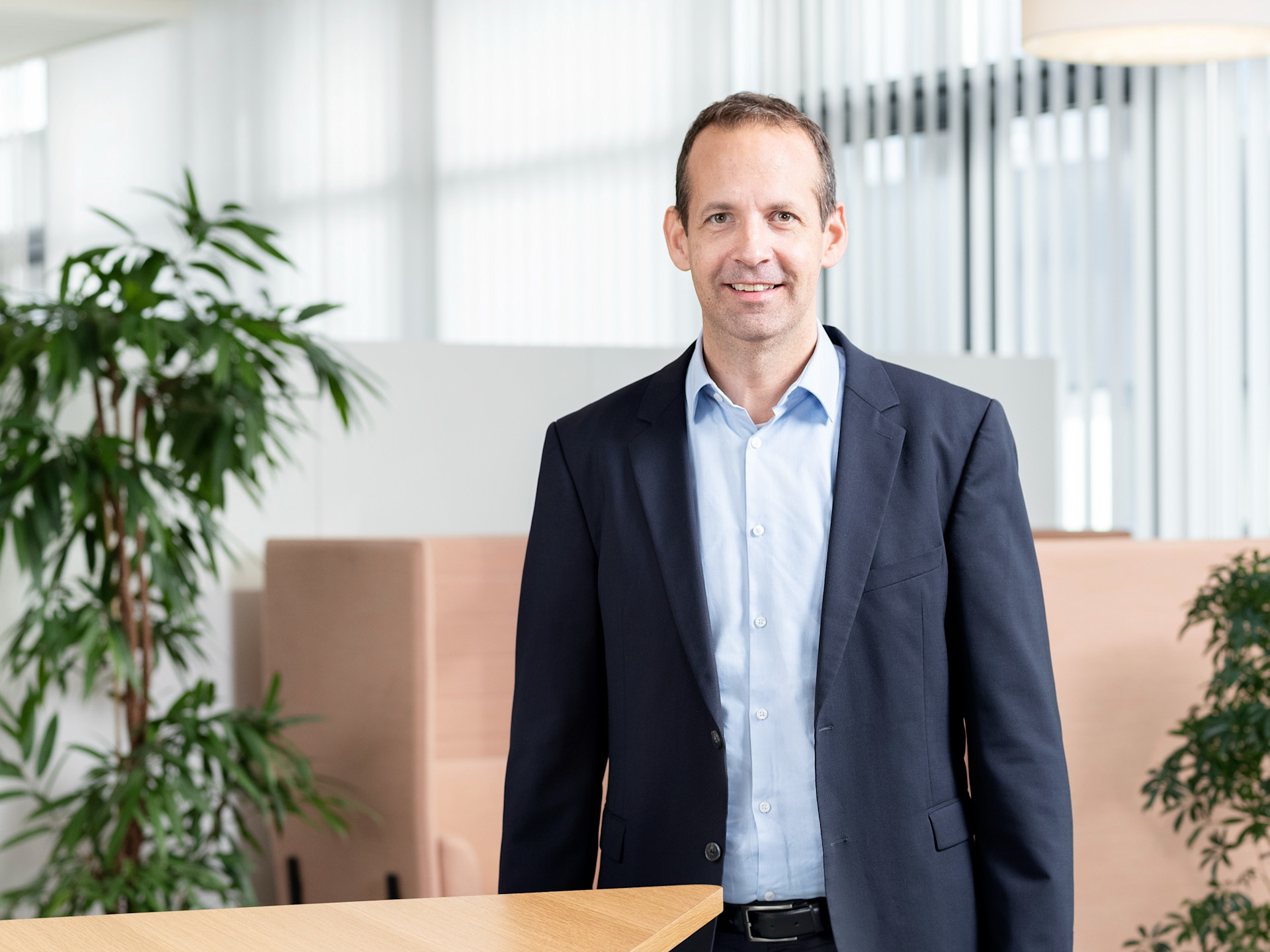 Roger Reinhard steht in Business Kleidung in einem modernen Büro mit Pflanzen, hellem Licht und klaren Linien im Hintergrund.