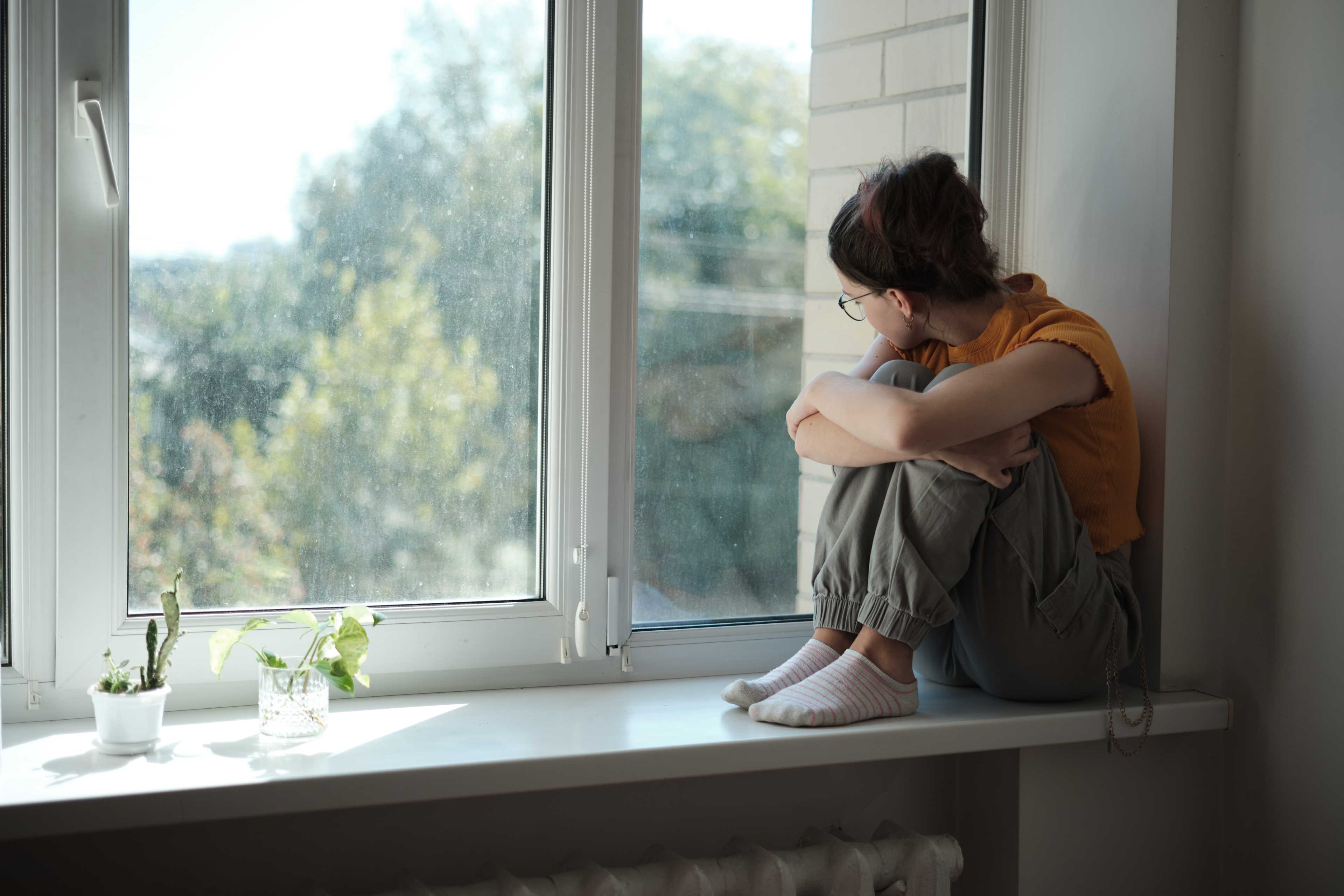 Una ragazza siede da sola sul davanzale di casa, abbraccia le ginocchia e guarda fuori dalla finestra.
