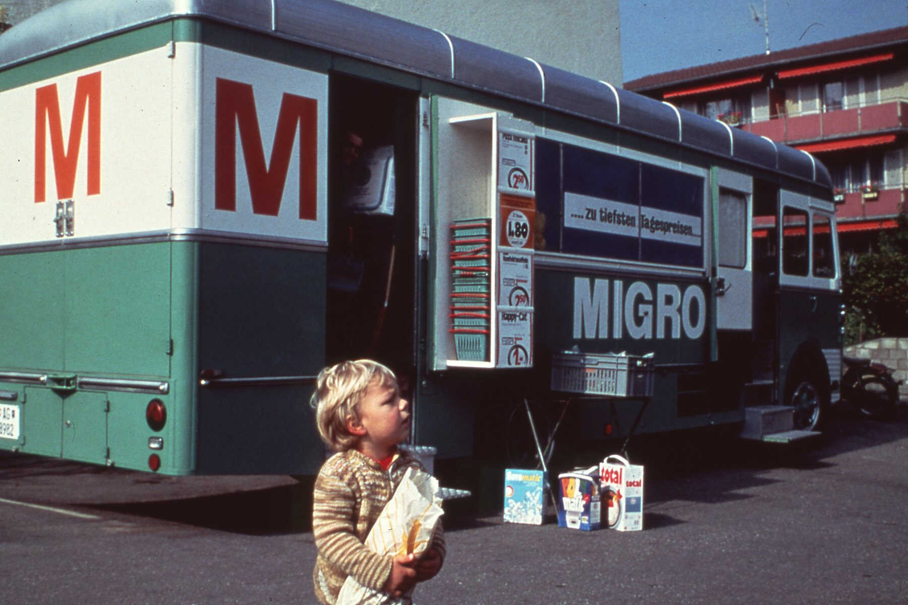 Un enfant avec un pain devant le chariot de vente Migros