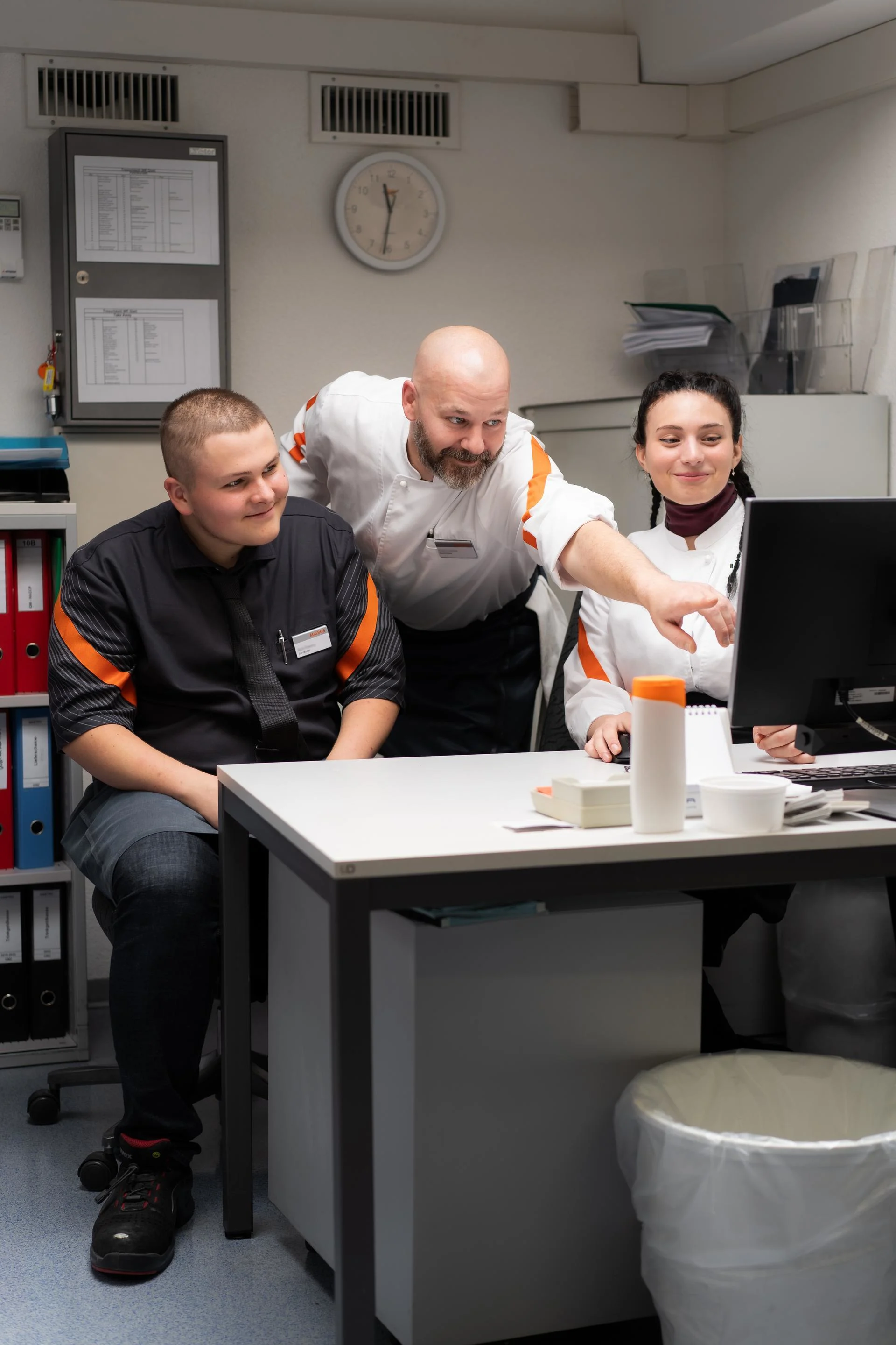 A supervisor explains something to two Migros restaurant apprentices at a desk.