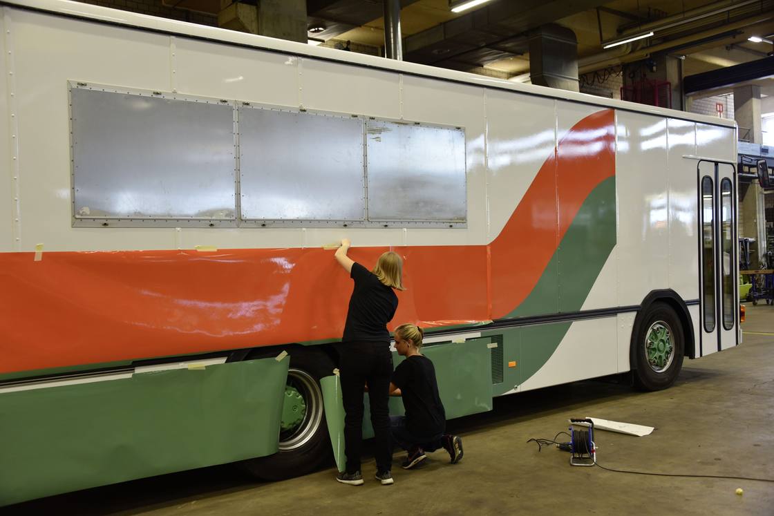 Zwei Personen kleben eine Folie mit orange-grünen Doppelstreifen auf einen Bus