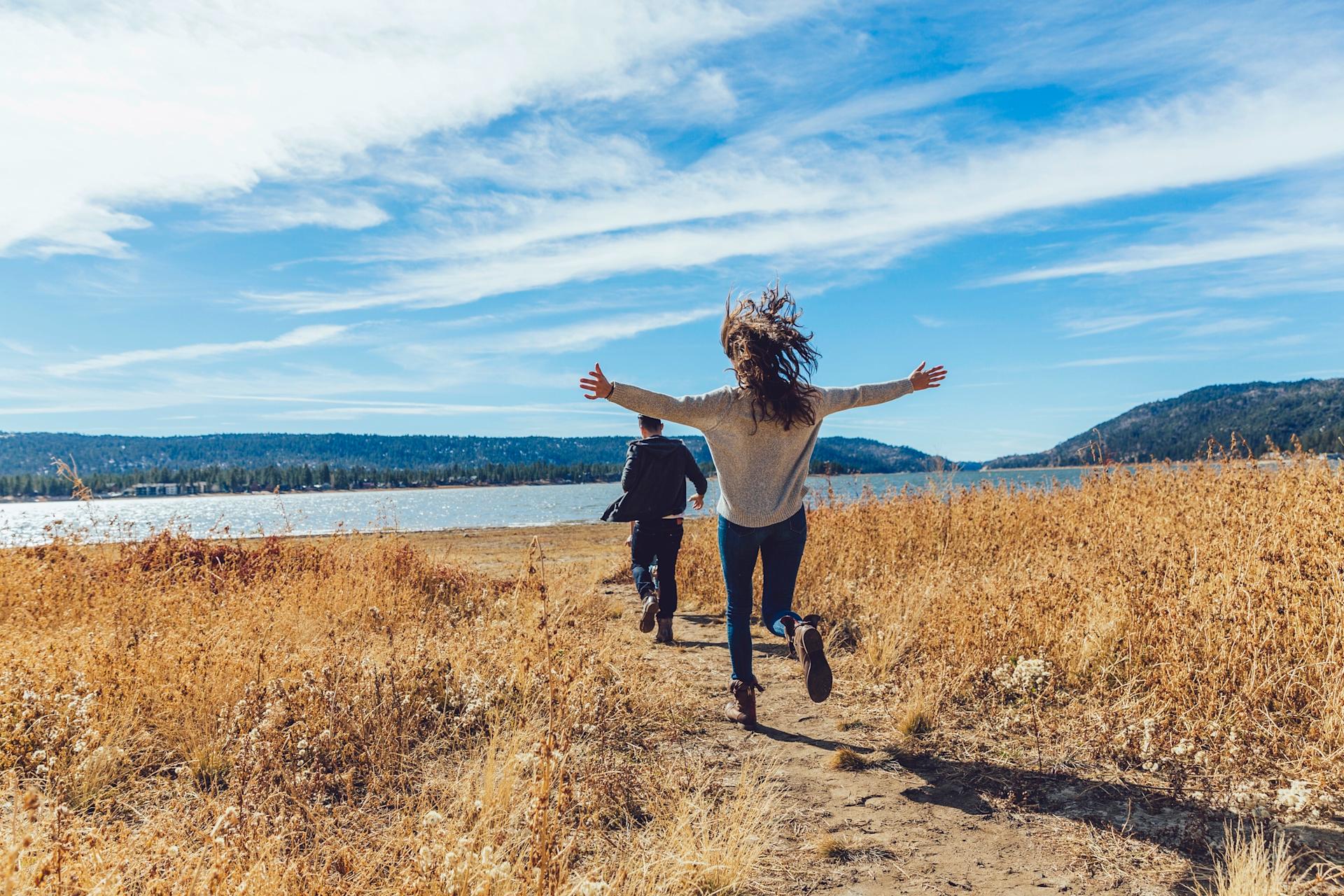 Two people walk happily along a path beside a lake in sunny weather, surrounded by golden grasses and a picturesque mountain landscape in the background.