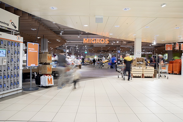 Eingangsbereich eines Migros-Supermarkts mit Einkaufswagen, Kundinnen und Kunden sowie dem leuchtenden Migros-Schriftzug im Hintergrund.