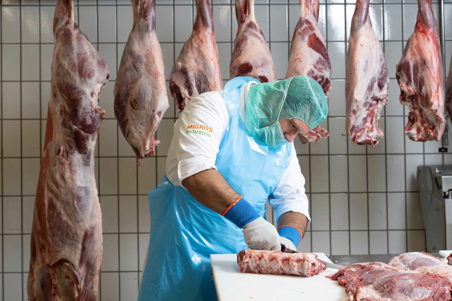 Marco Eisenlohr trägt einen Schutzanzug und schneidet ein Stück Fleisch entzwei. Im Hintergrund hängen ganze Fleischstücke an Haken.