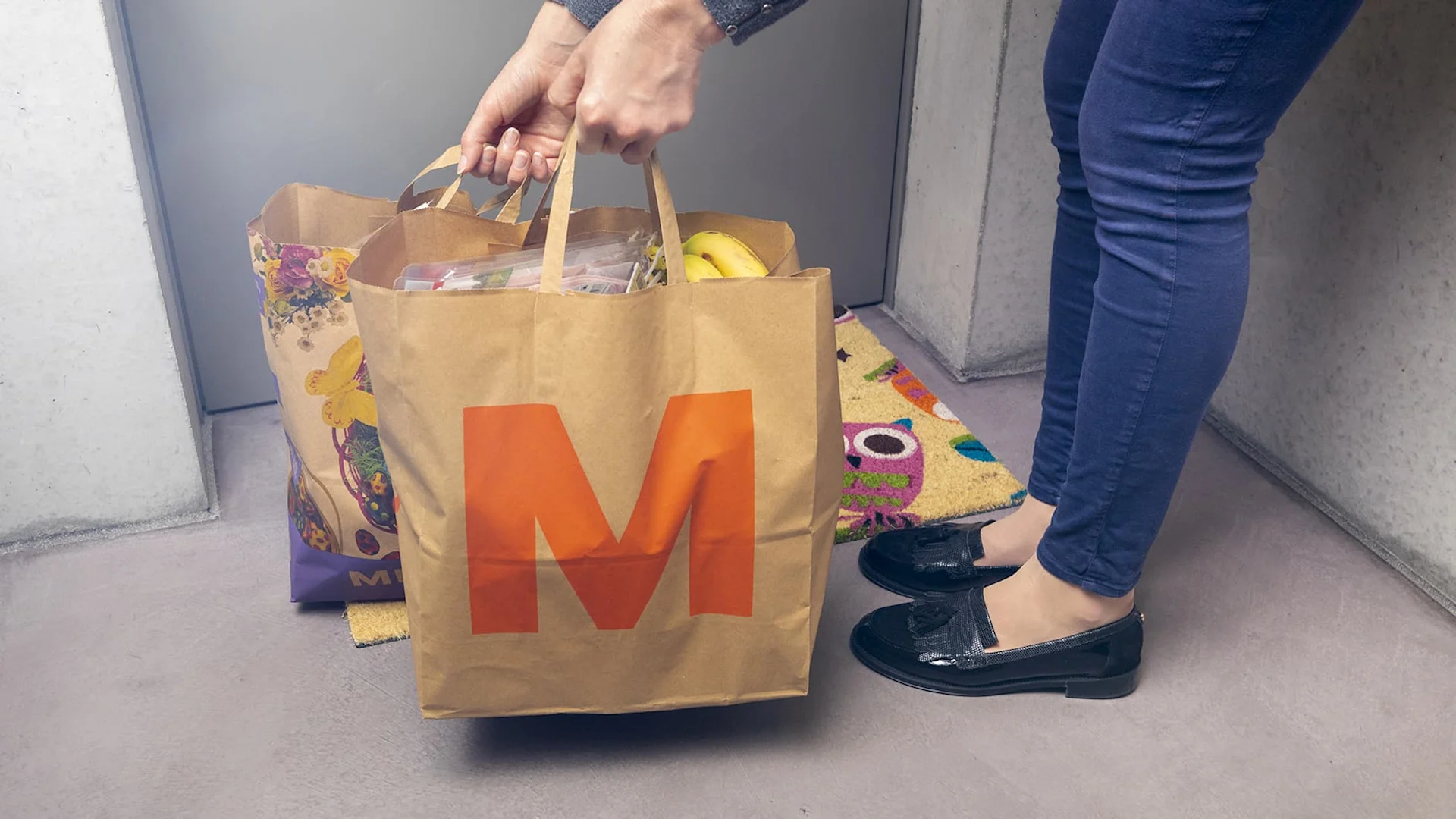 Eine Person stellt zwei Migros-Taschen vor einer Tür ab