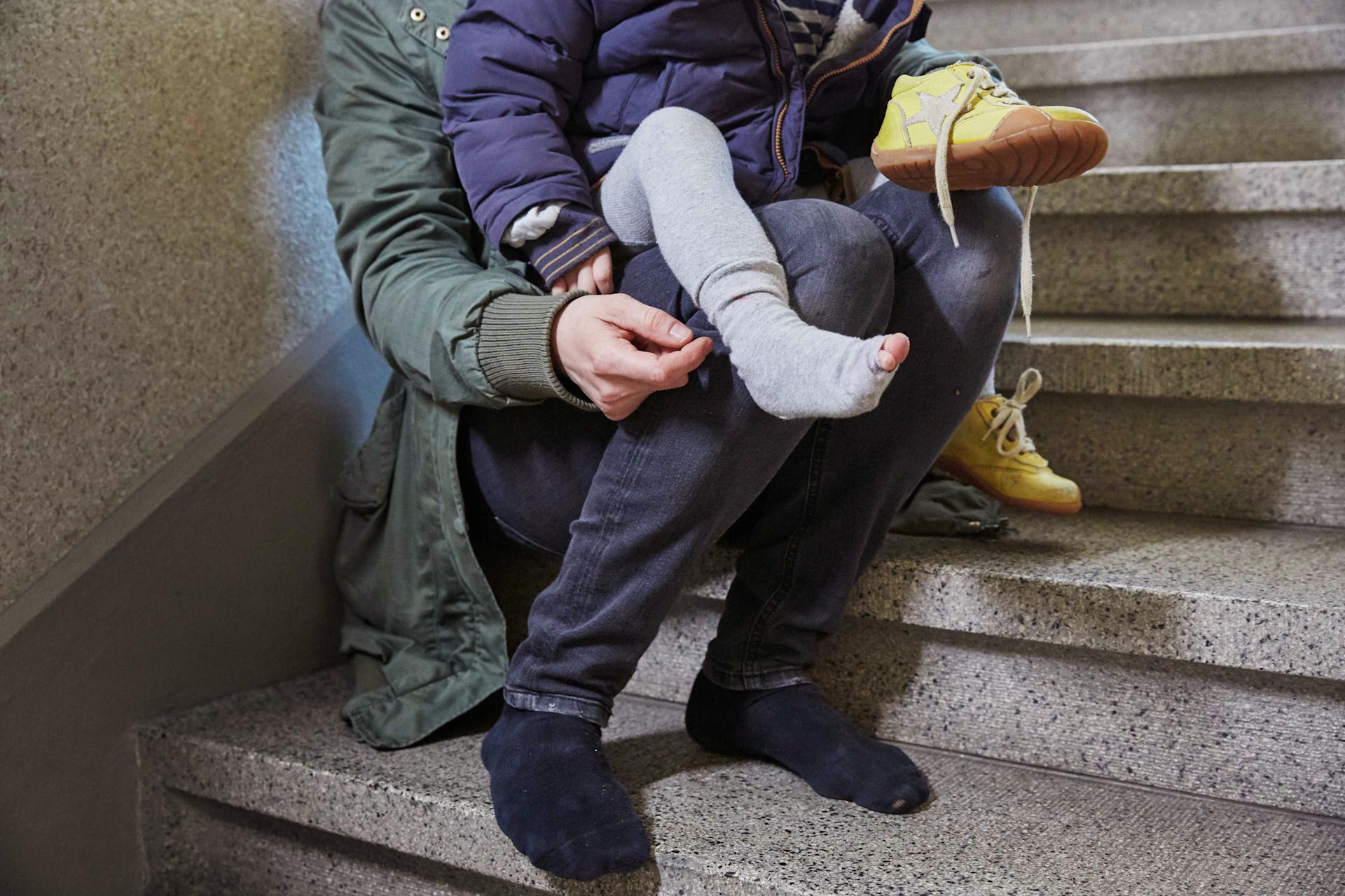 Eine Mutter sitzt im Treppenhaus und zieht ihrem Kind, in dessen Strumpf sich ein Loch befindet, die Schuhe an.