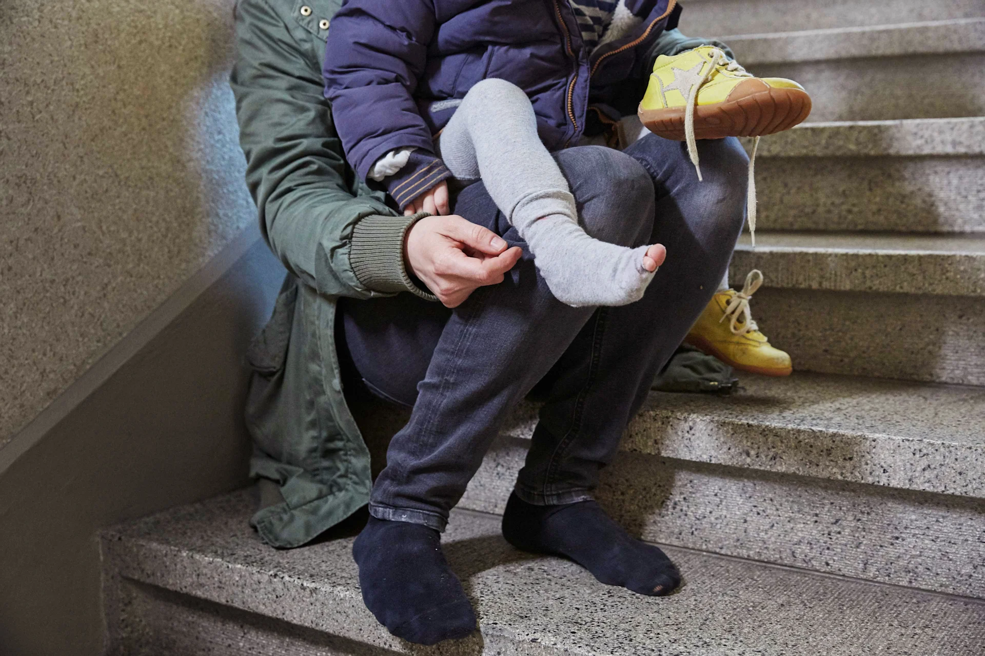 Eine Mutter sitzt im Treppenhaus und zieht ihrem Kind, in dessen Strumpf sich ein Loch befindet, die Schuhe an.