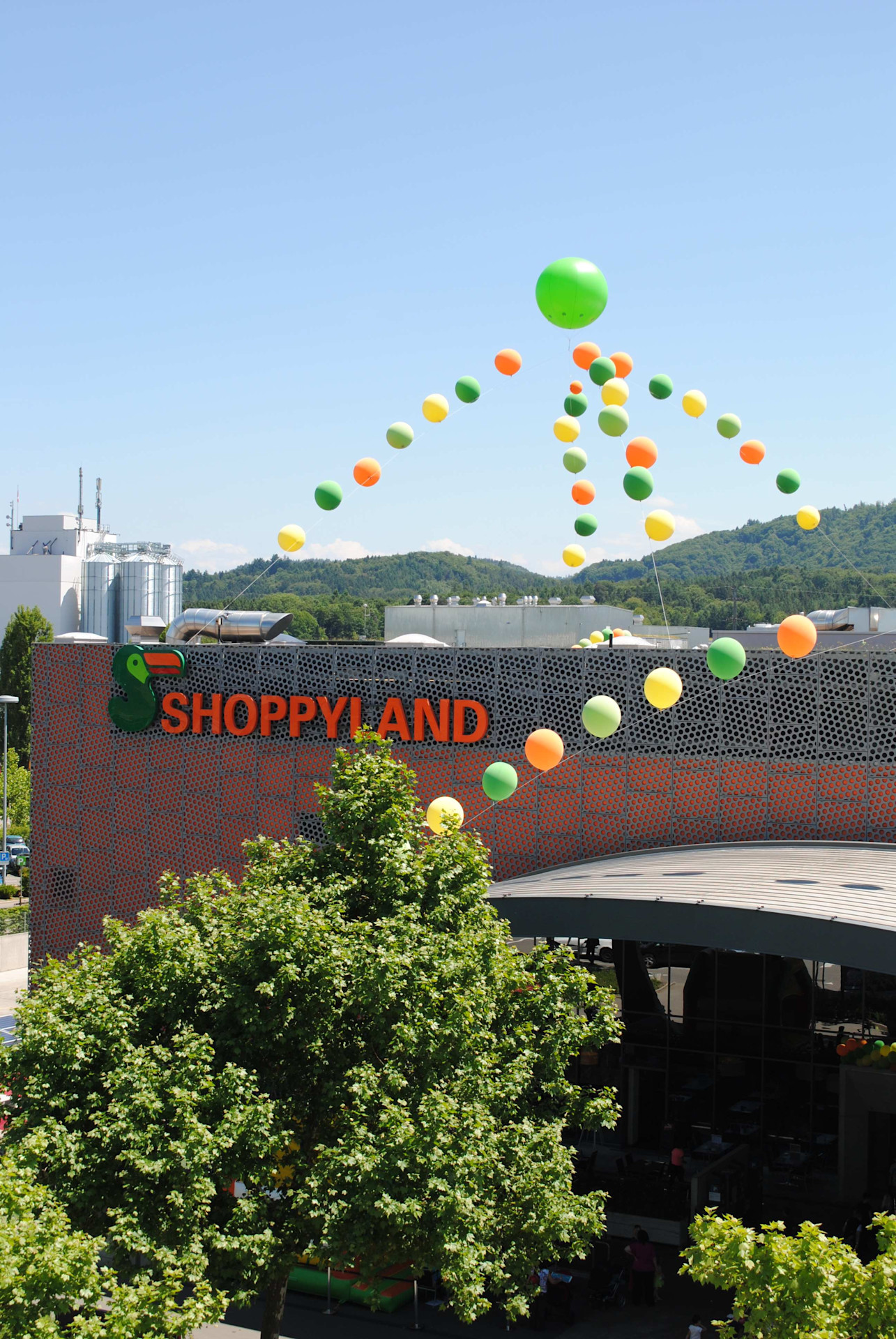 Aussenansicht des Einkaufszentrums Shoppyland mit farbigen Ballons am klaren Himmel und umgebender grüner Landschaft.