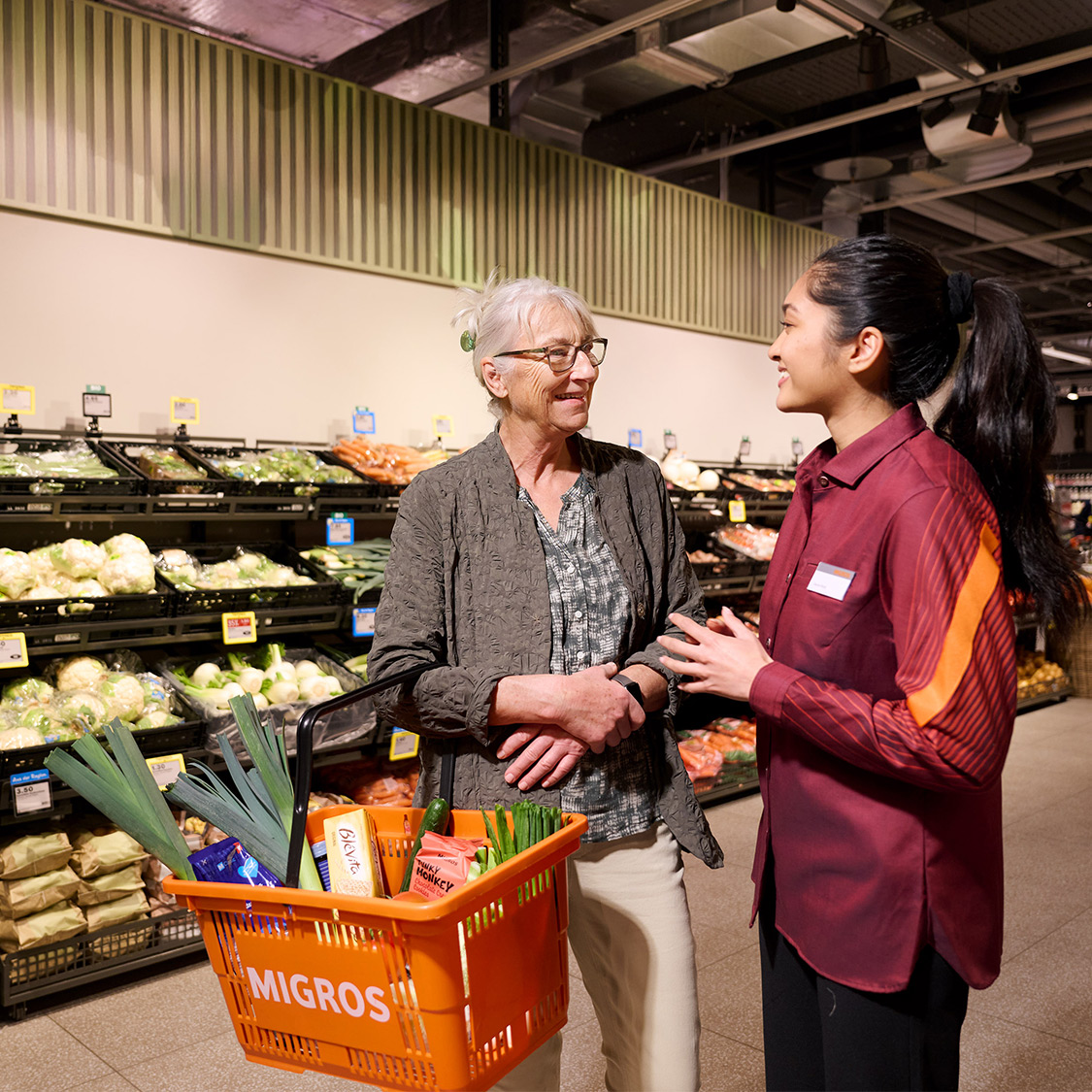 Collaboratrice Migros avec une cliente en magasin qui porte un panier de produits