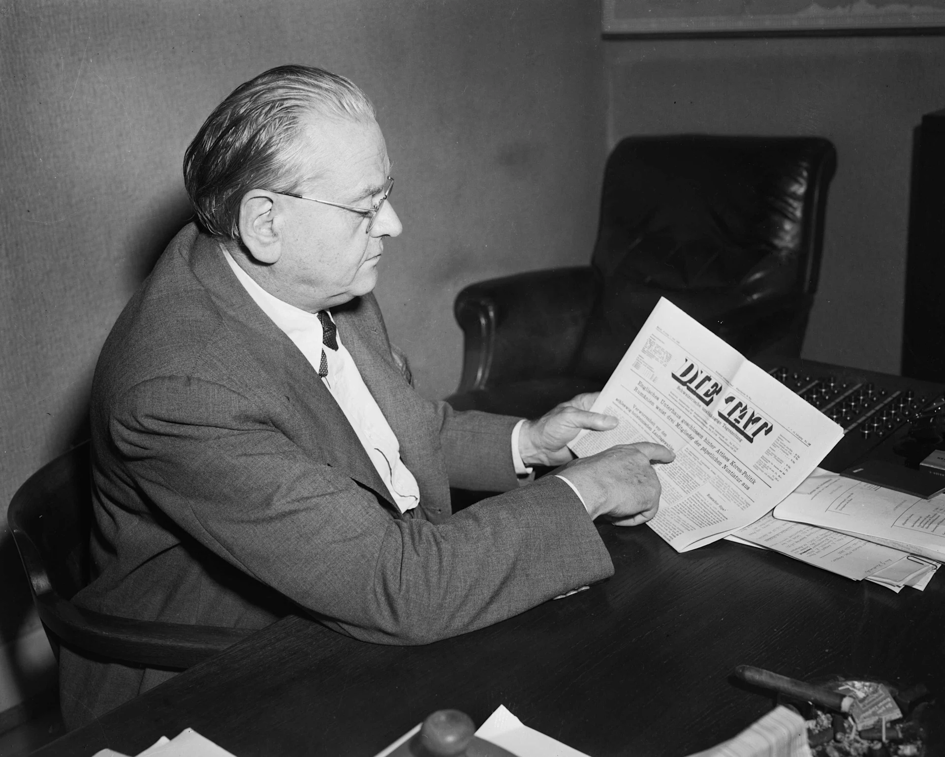 A black-and-white photo of Gottlieb Duttweiler reading the newspaper "Die Tat"