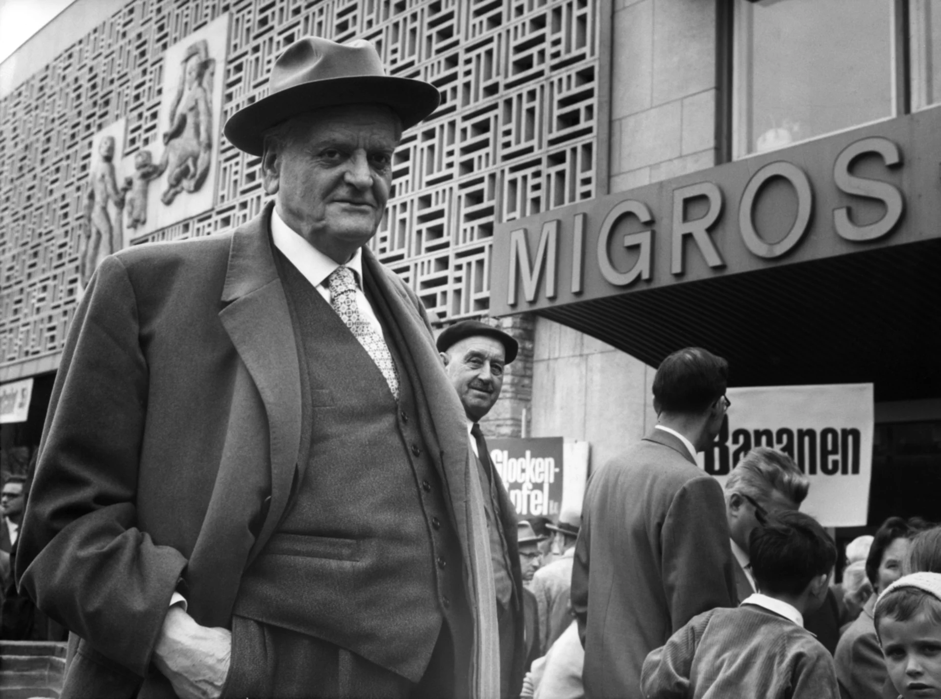 A black-and-white photograph of Gottlieb Duttweiler standing in front of a Migros store, looking into the camera.