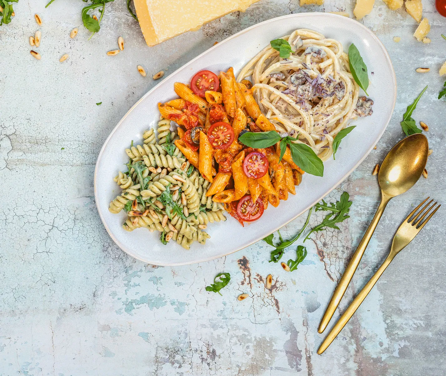 Teller mit drei verschiedenen Pastasorten: grüne Fusilli mit Kräutern, Penne in roter Tomatensauce mit Cherrytomaten und Basilikum, sowie Spaghetti in heller Rahmsauce mit Zwiebeln. Pinienkerne auf dem Tisch.