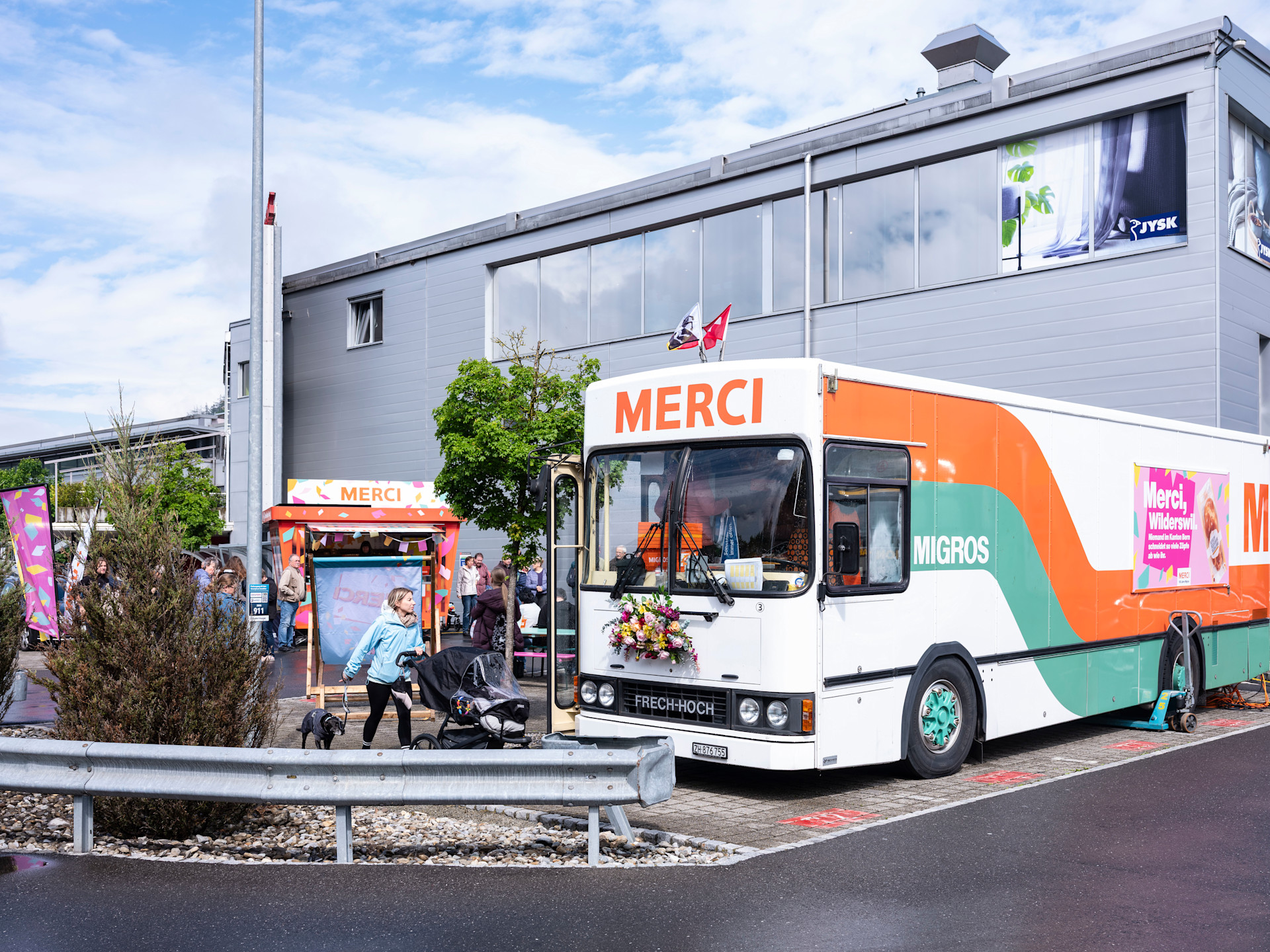 Un bus Migros arborant un grand «Merci» est garé devant un bâtiment, à côté duquel se trouvent un petit stand d'information et diverses affiches. Scène d'une action ou d'une tournée devant un bâtiment d'entreprise gris.