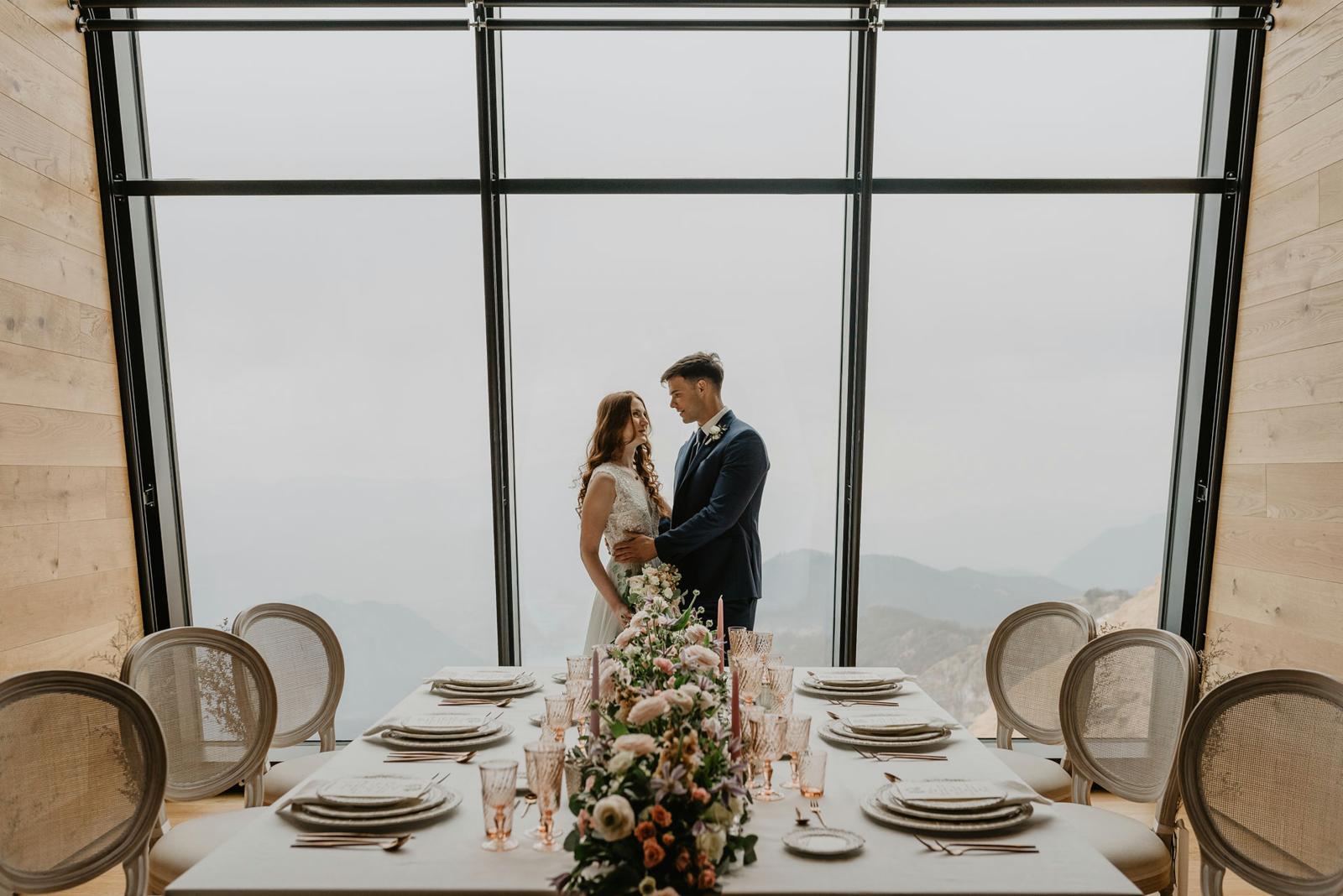 A wedding couple poses in front of the window with a view of the mountains and a laid table with wedding decorations in the foreground.