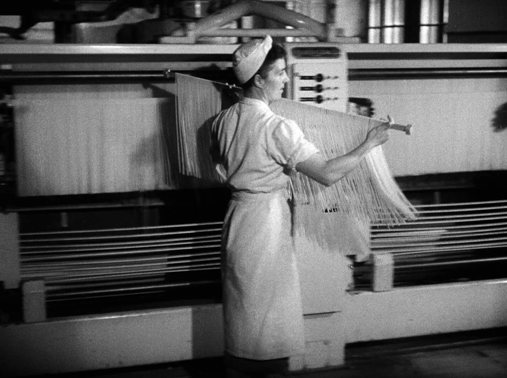 A woman in a factory carries a rod over which pasta hangs.