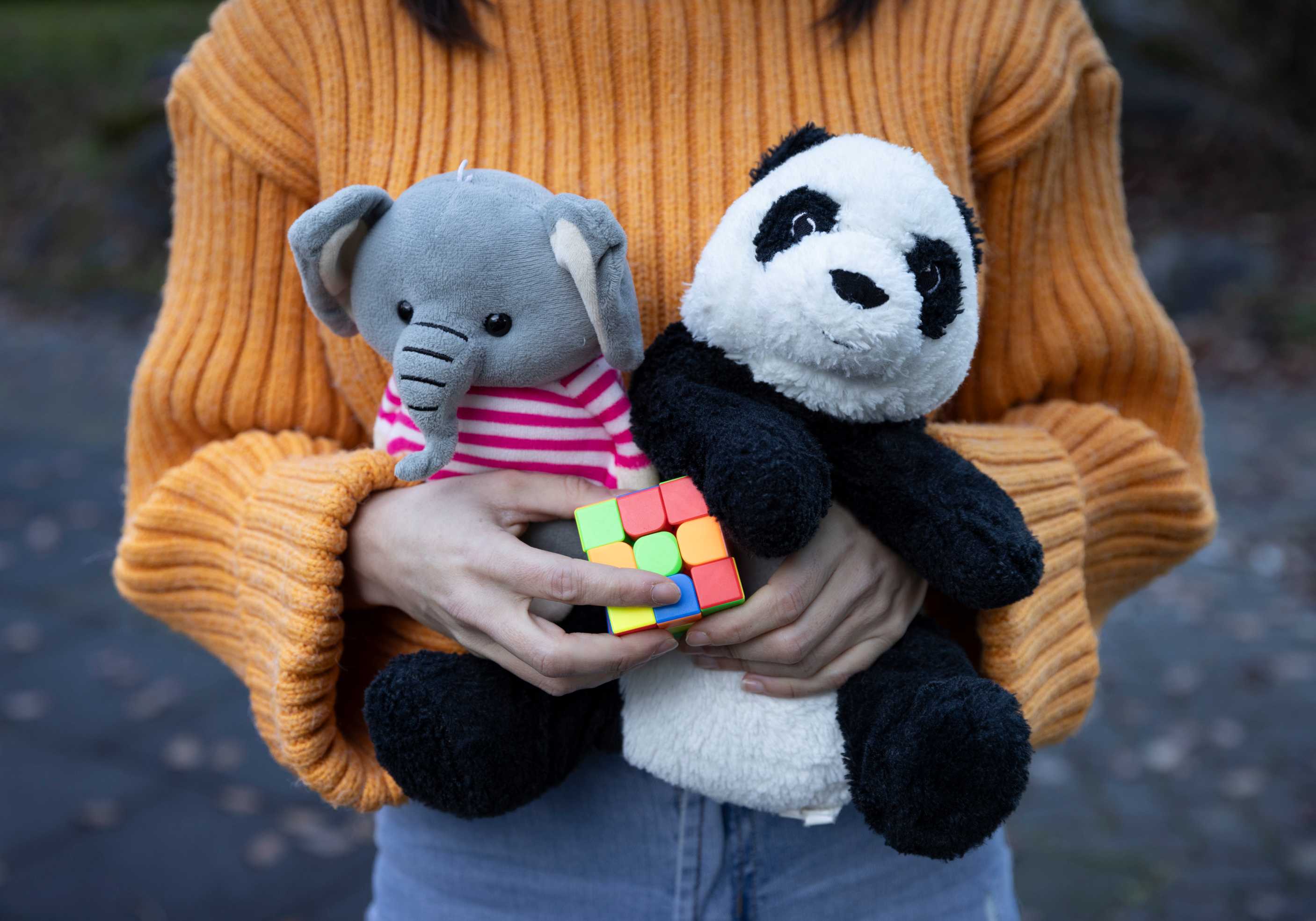 Eine Frau im orangen Pullover hält zwei Plüschtiere und einen Würfel im Arm.