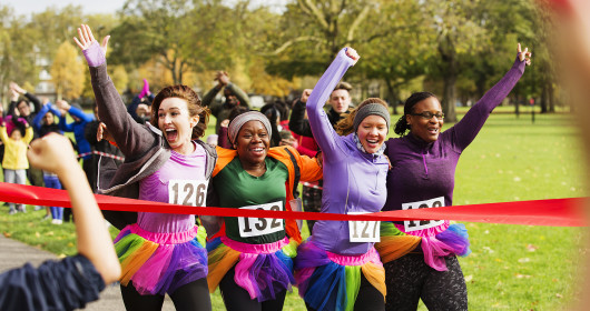 Enthusiastic female runners in tutus crossing charity run finish line in park, celebrating 