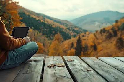 Person reading e-reader on wooden deck overlooking autumn mountains with orange and green foliage under cloudy sky.