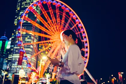Smiling young woman using smartphone in city, against illuminated ferris wheel and city skyline at night