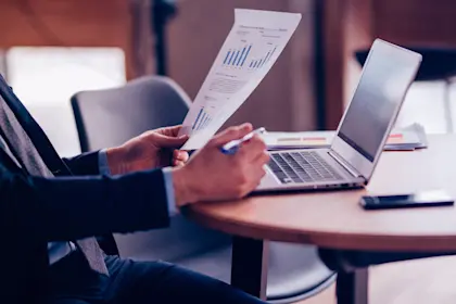 Businessman sitting at table reviewing document with laptop opened behind it