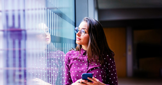 Shot of a young female business professional with a cell phone standing by a glass wall in office 
