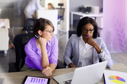 Two businesswomen are in the office using laptop, digital tablet and chart papers while working together.