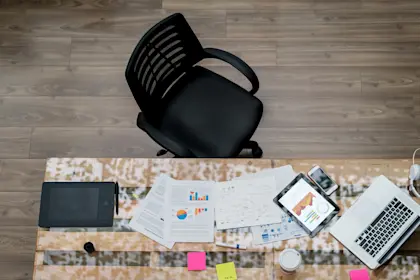 Overhead view of papers and laptop on top of desk with empty chair slightly pulled out.
