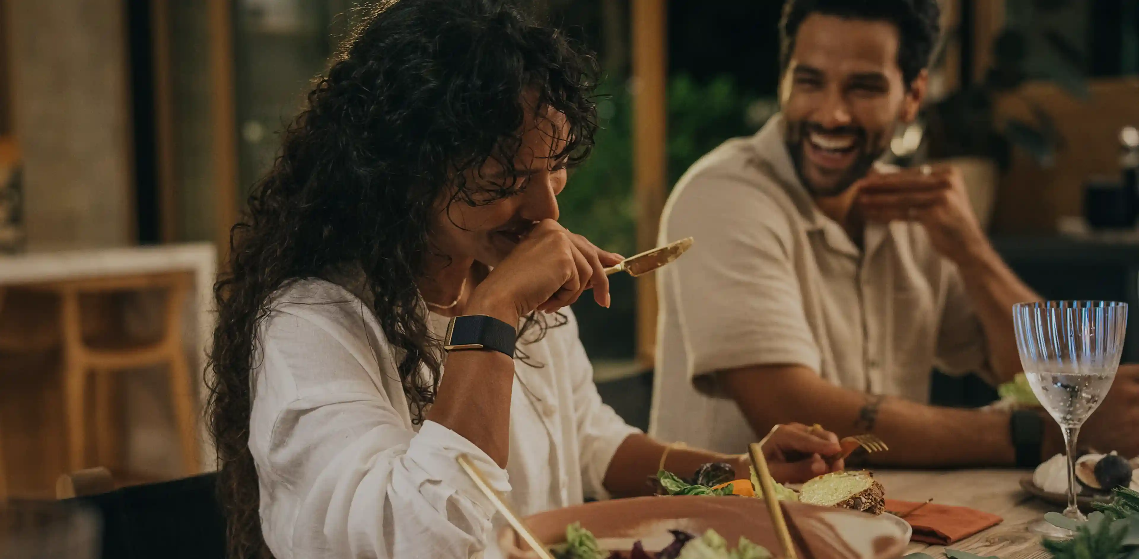 Evening dinner scene with friends; a woman wearing a WHOOP 5.0 band laughs at the table while serving salad, illustrating everyday wear that tracks heart rate, HRV, and recovery during social moments.