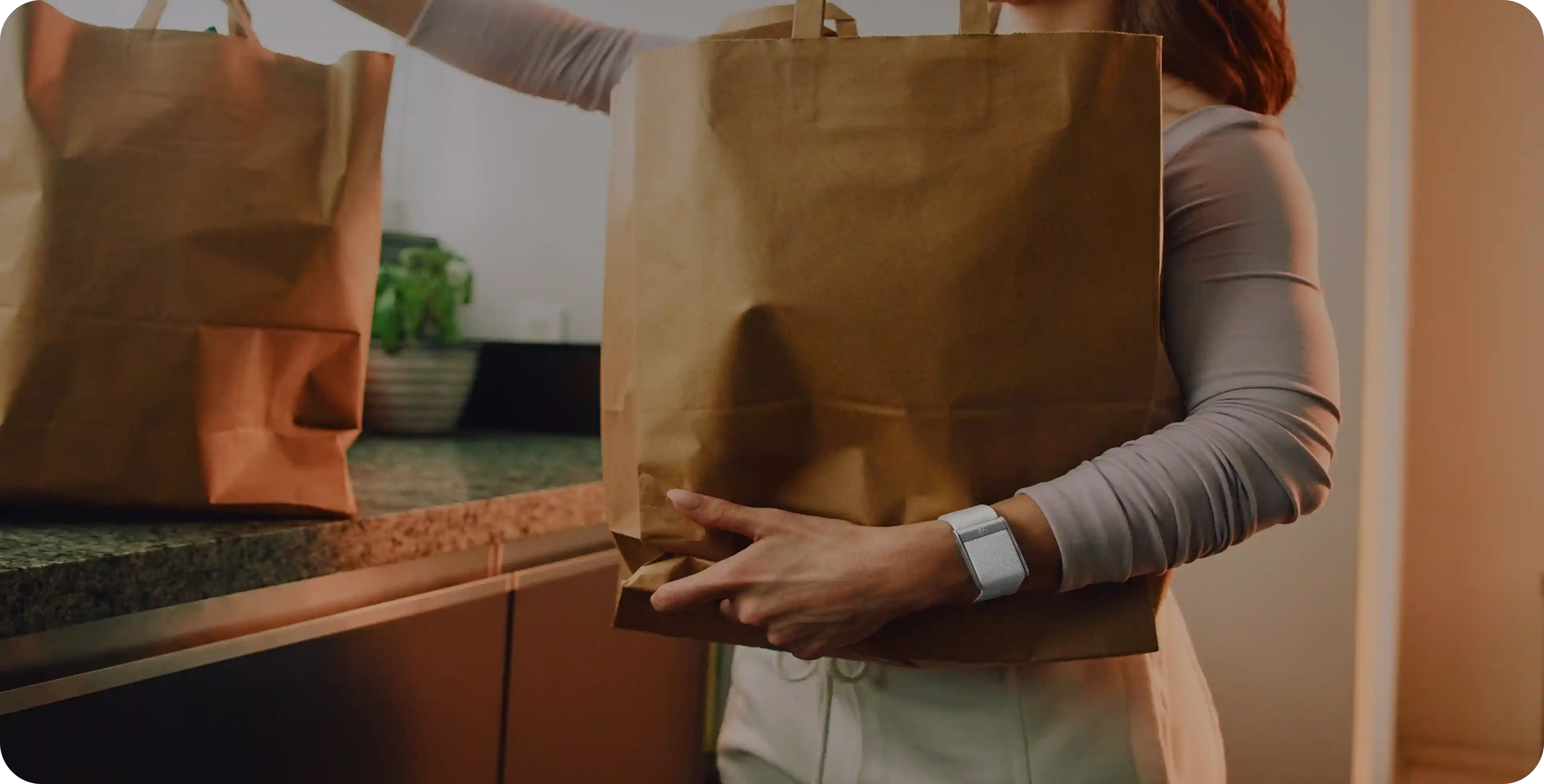 Person wearing a WHOOP 5.0 band carrying a paper grocery bag in the kitchen after a store run, highlighting everyday wear that tracks heart rate, strain, and recovery during daily tasks.