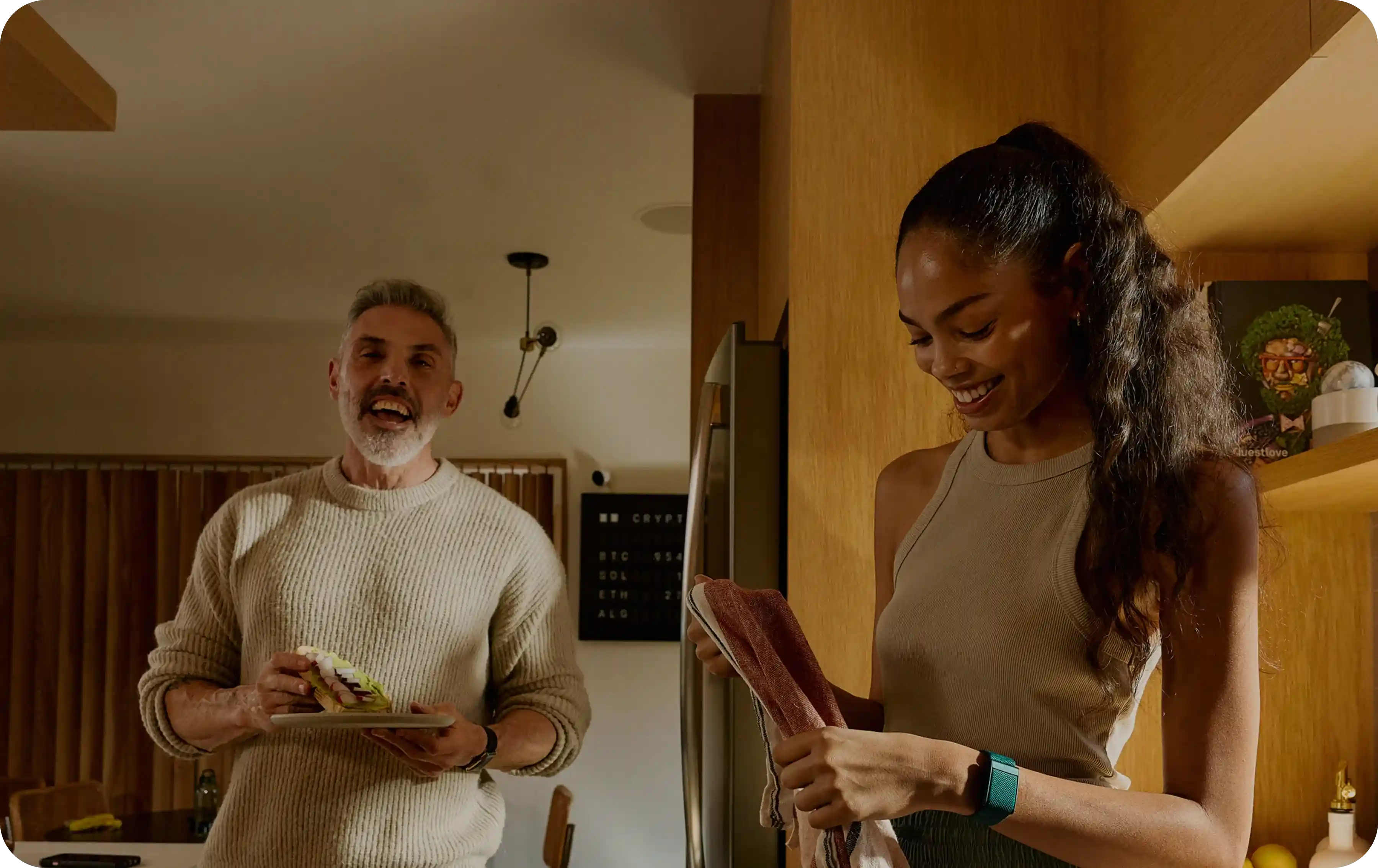 In a warm kitchen, a woman wearing a WHOOP 5.0 band dries dishes while chatting with a companion, illustrating everyday wear that continuously tracks heart rate, strain, and recovery during daily life.