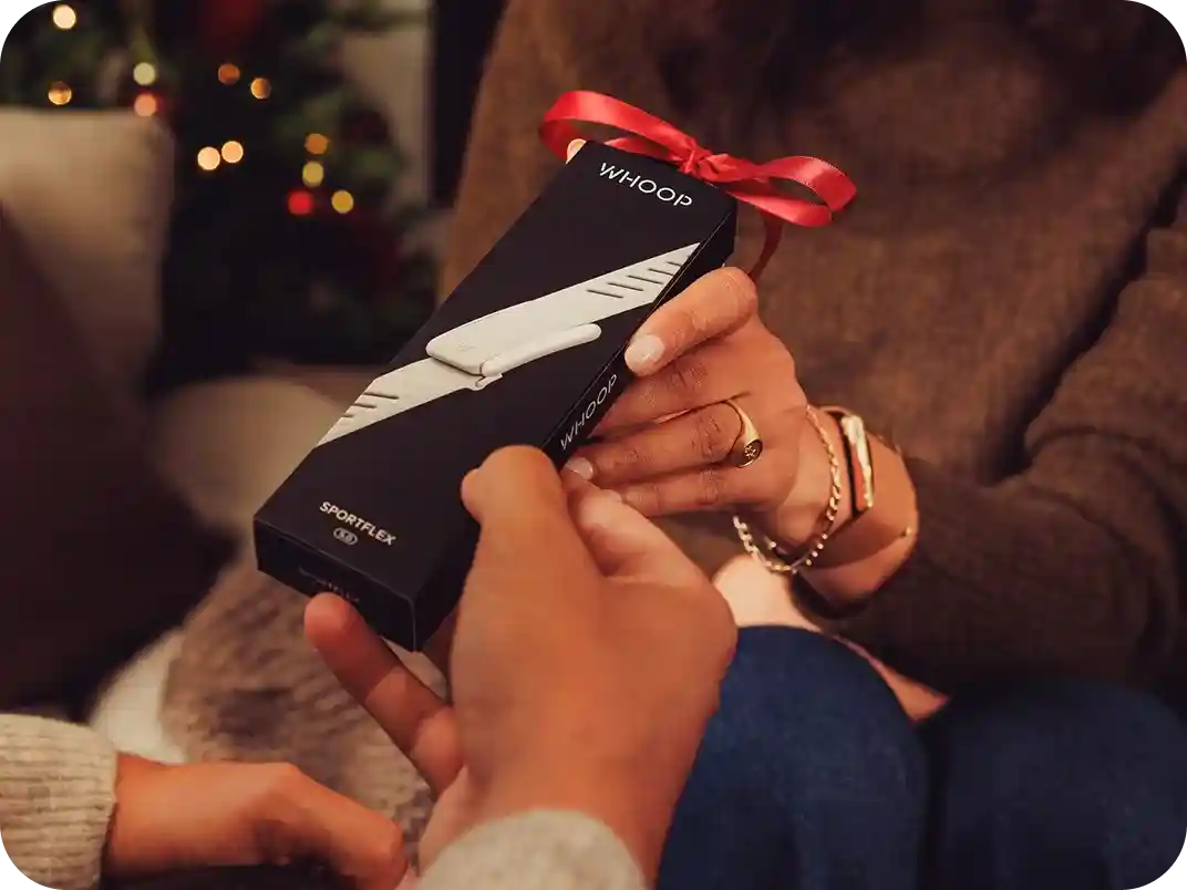 Close-up of two people exchanging a WHOOP gift box tied with a red ribbon in a cozy living room with tree lights, showcasing the ready-to-gift packaging for the WHOOP 5.0 band.
