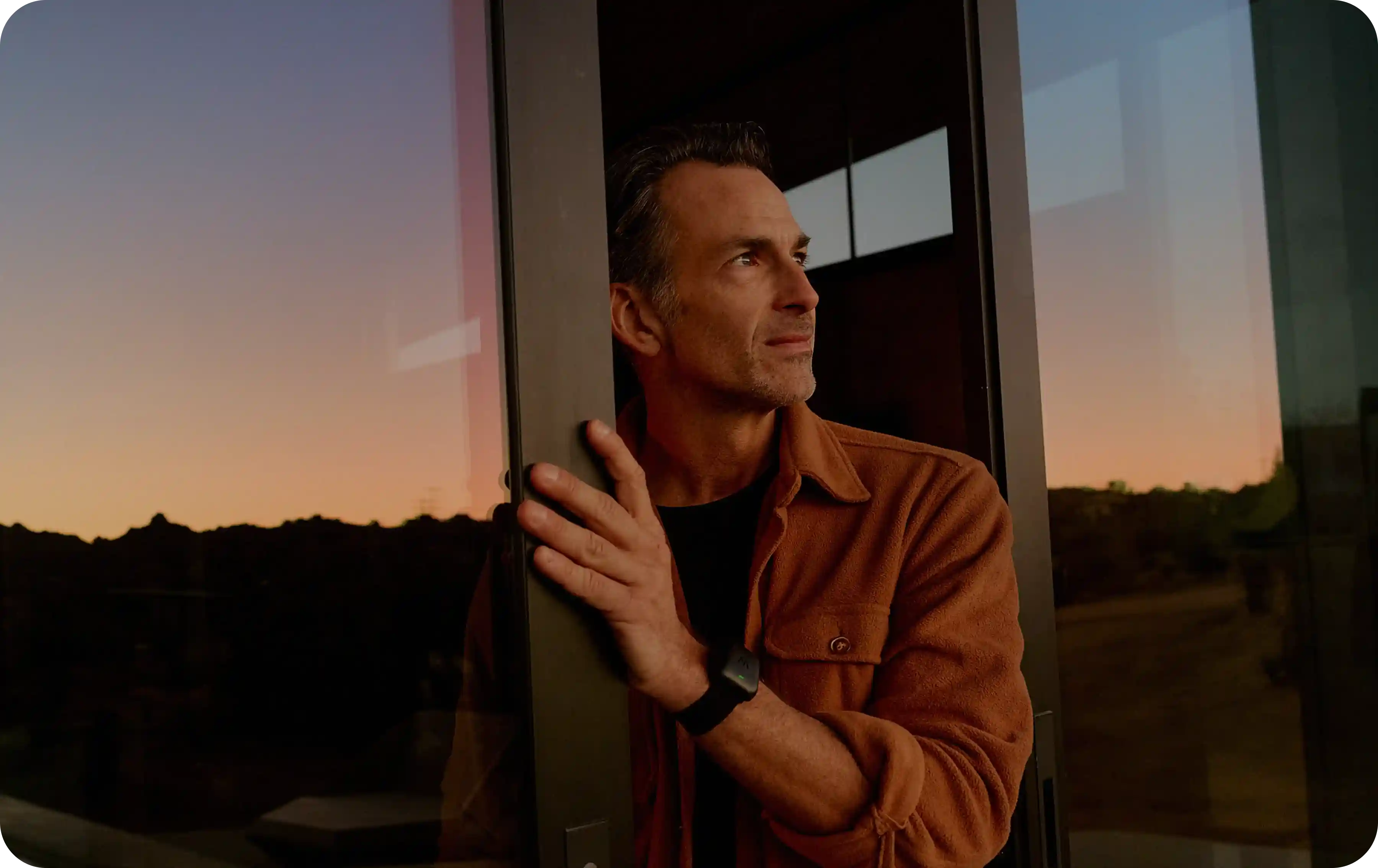 Man wearing a WHOOP 5.0 band standing in a glass doorway at sunset, warm reflections on the window, highlighting everyday wear for continuous heart rate, recovery, and sleep tracking.