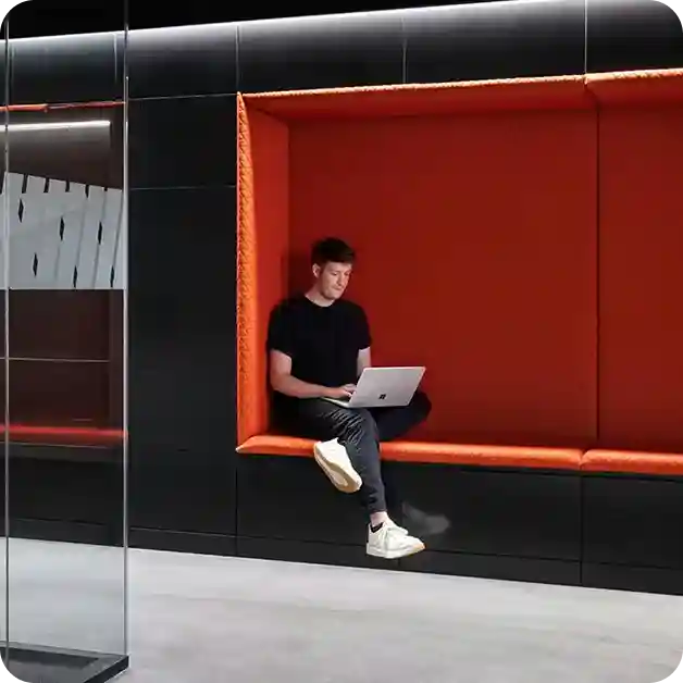 Man working on a laptop in a modern orange seating nook inside the WHOOP office, highlighting collaborative workspace design.