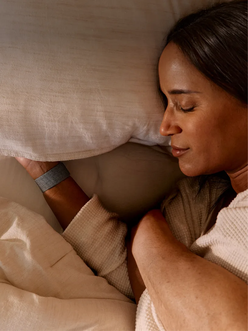 Woman with dark hair resting peacefully on beige pillow wearing a fitness tracker, eyes closed in serene sleep position.