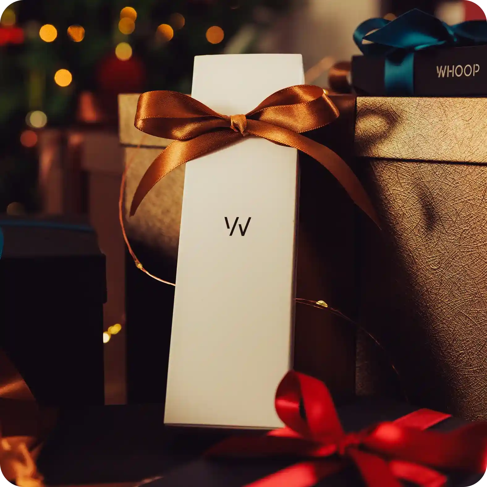 Close-up of a tall white WHOOP gift box tied with a satin copper bow, set among wrapped presents and warm tree lights, highlighting ready-to-gift packaging for the WHOOP 5.0 band or accessories.