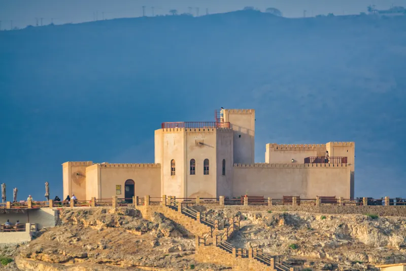 Forteresse beige sur une falaise rocheuse, ciel bleu et montagnes en arrière-plan, Dhofar, Salalah, Oman.