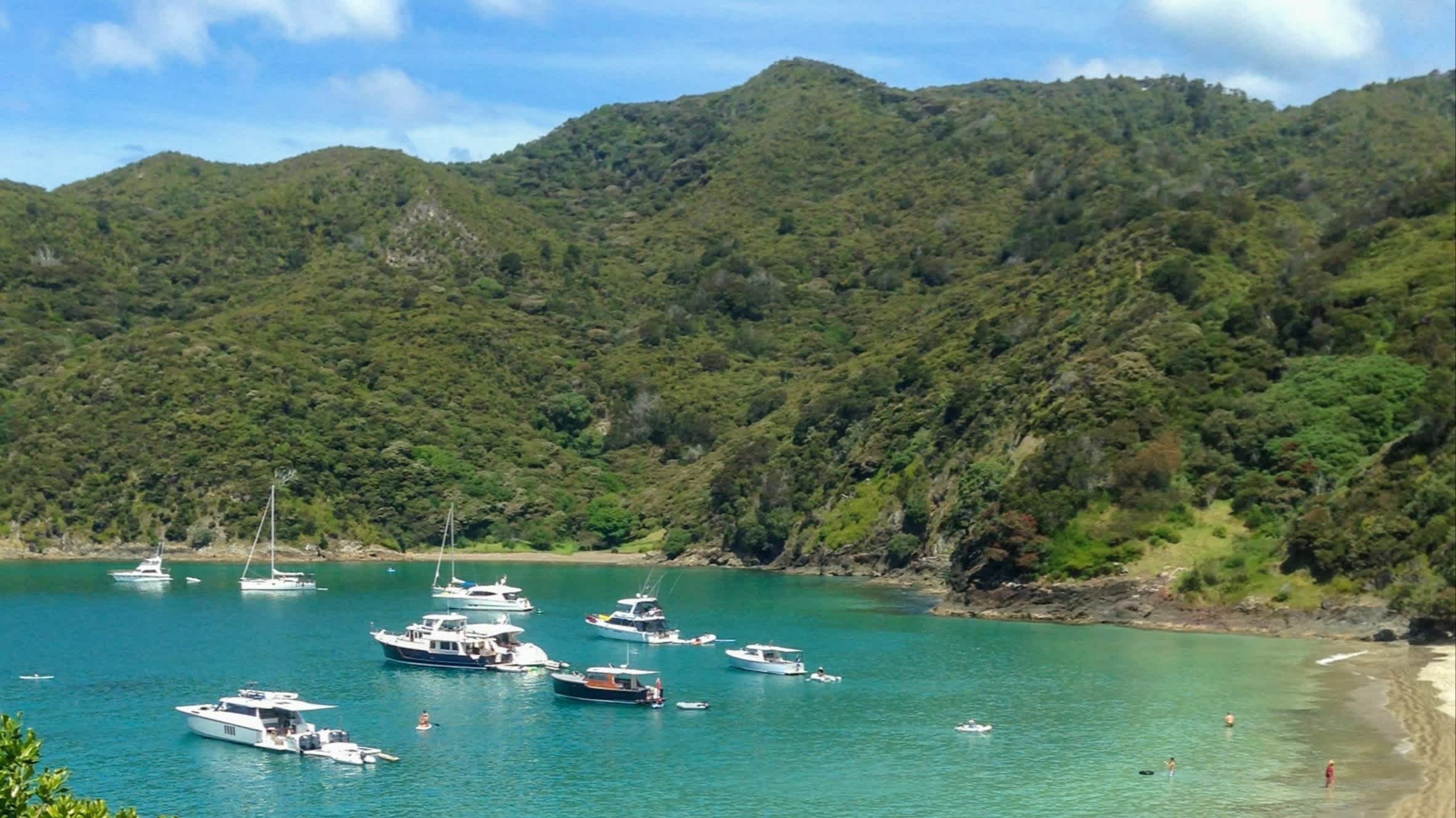 Blick auf die Bucht Oke Bay, Bay of Islands, Neuseeland, bei Sonnenschein und mit Booten im Wasser sowie grünen Hügeln und dem türkisfarbenem Meer.

