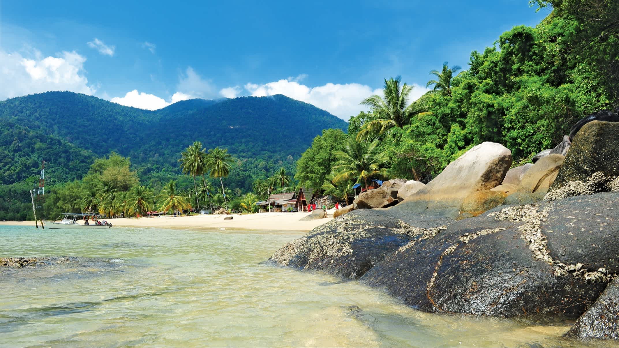 Felsen am weißen Sandstrand von Pulau Tioman, Malaysia mit Blick auf die tropische Umgebung und mit kleinen Strandhäusern im Bild.