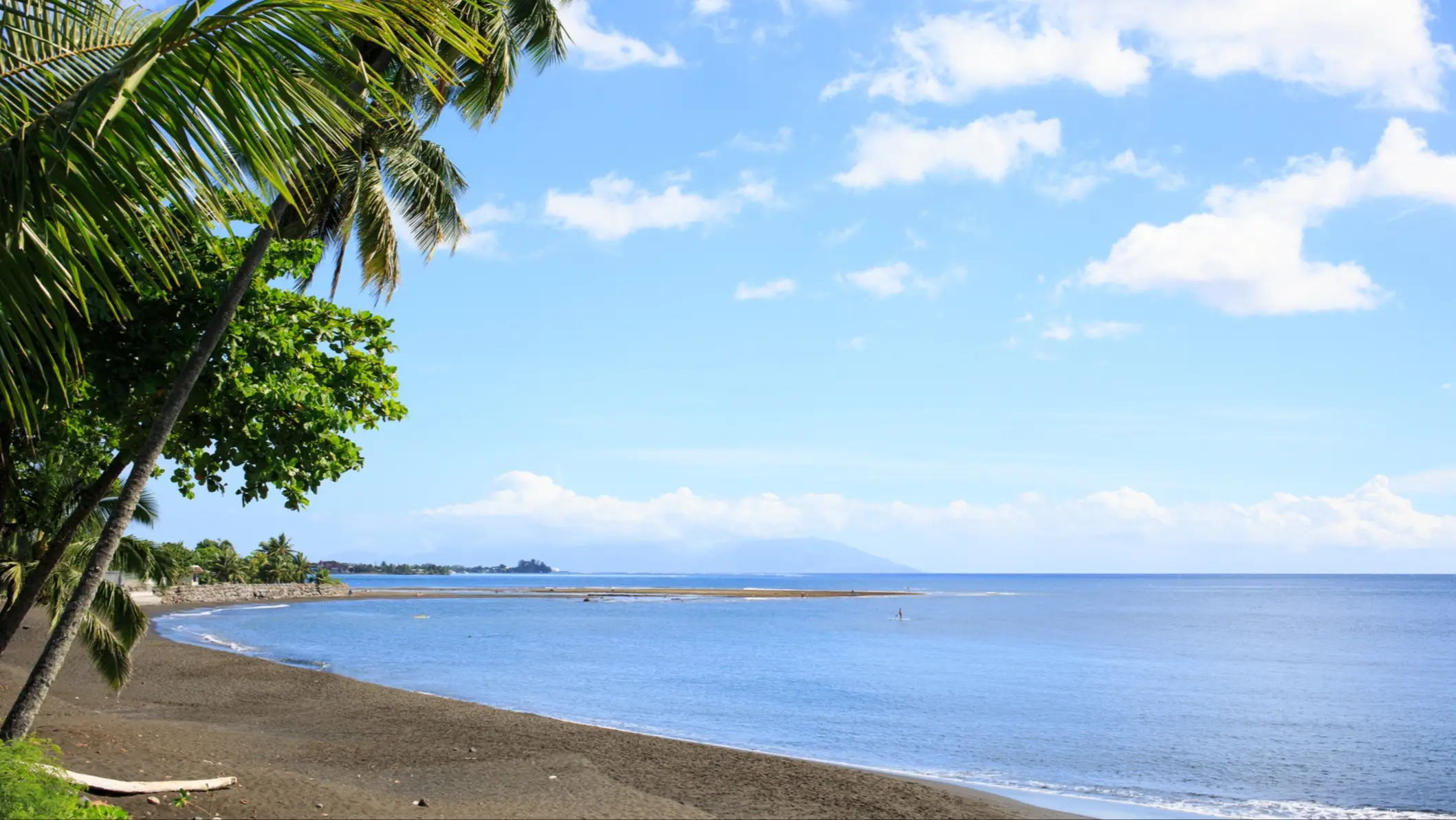 Plage tropicale avec sable noir, mer bleue calme, palmiers verts et ciel bleu parsemé de nuages blancs, Papeete.